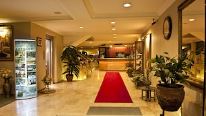 Un vestíbulo de hotel con alfombra roja, mostrador de recepción, plantas en macetas y un espejo grande.