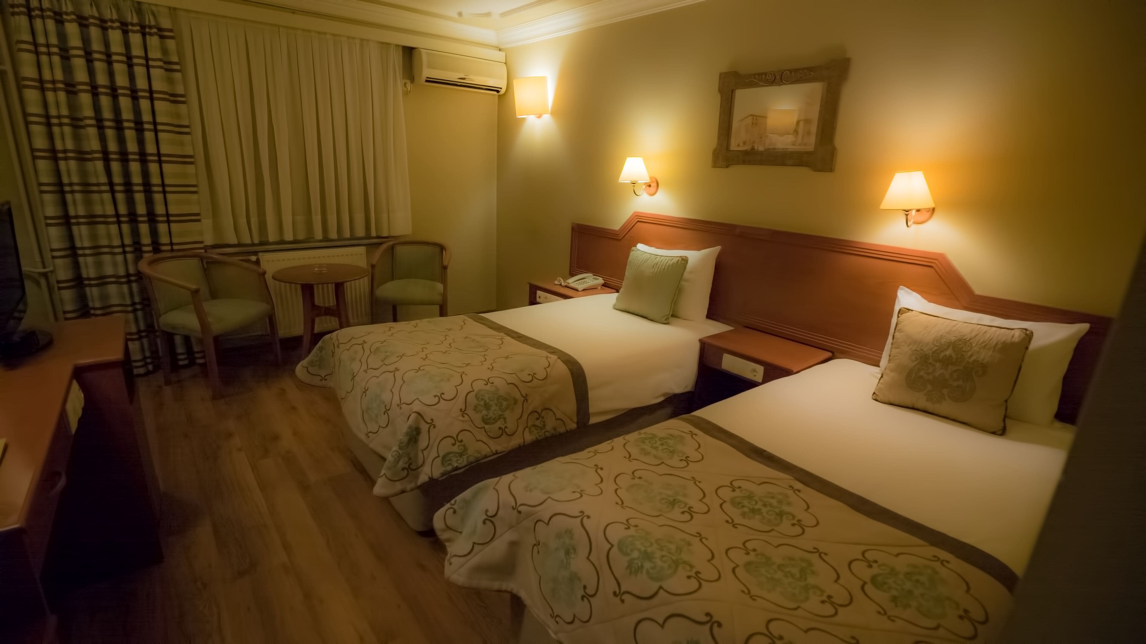 Ein Hotelzimmer mit zwei Betten, einem Schreibtisch und einem Stuhl. An den Wänden sind Lampen angebracht und es gibt eine Klimaanlage.