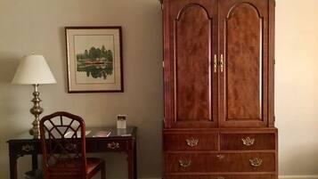 A wooden cabinet with two doors and multiple drawers, a wooden chair, a desk with a lamp, and a framed picture on the wall.