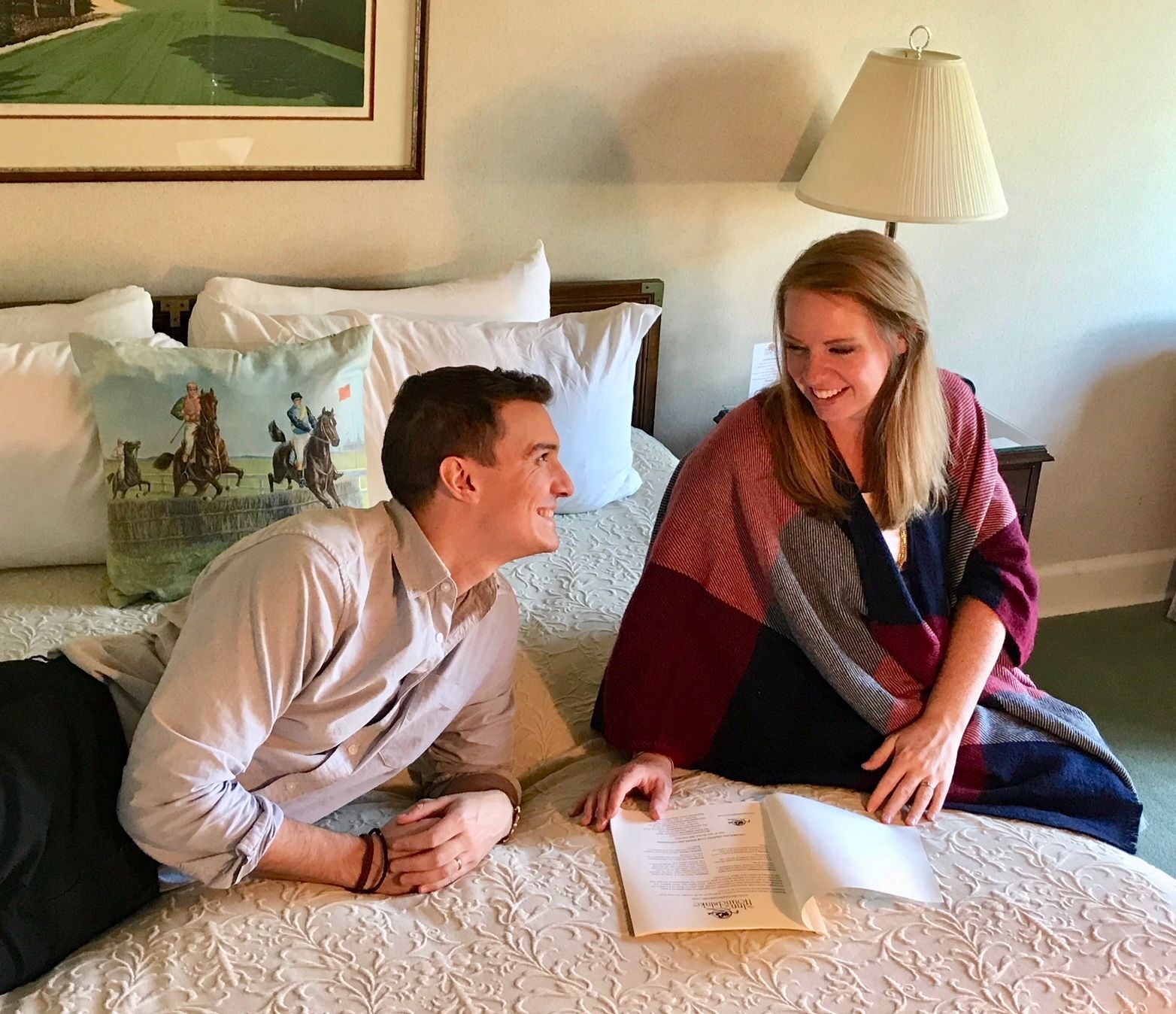 Una pareja sentada en una cama con un libro, en una habitación con un cuadro y una lámpara.