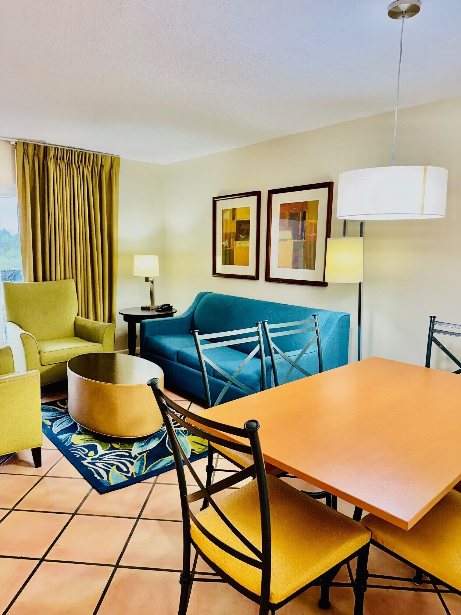 A modern living room with a blue sofa, yellow armchairs, a round coffee table, and a dining table with chairs. There are two framed artworks on the wall and a lamp on a side table.