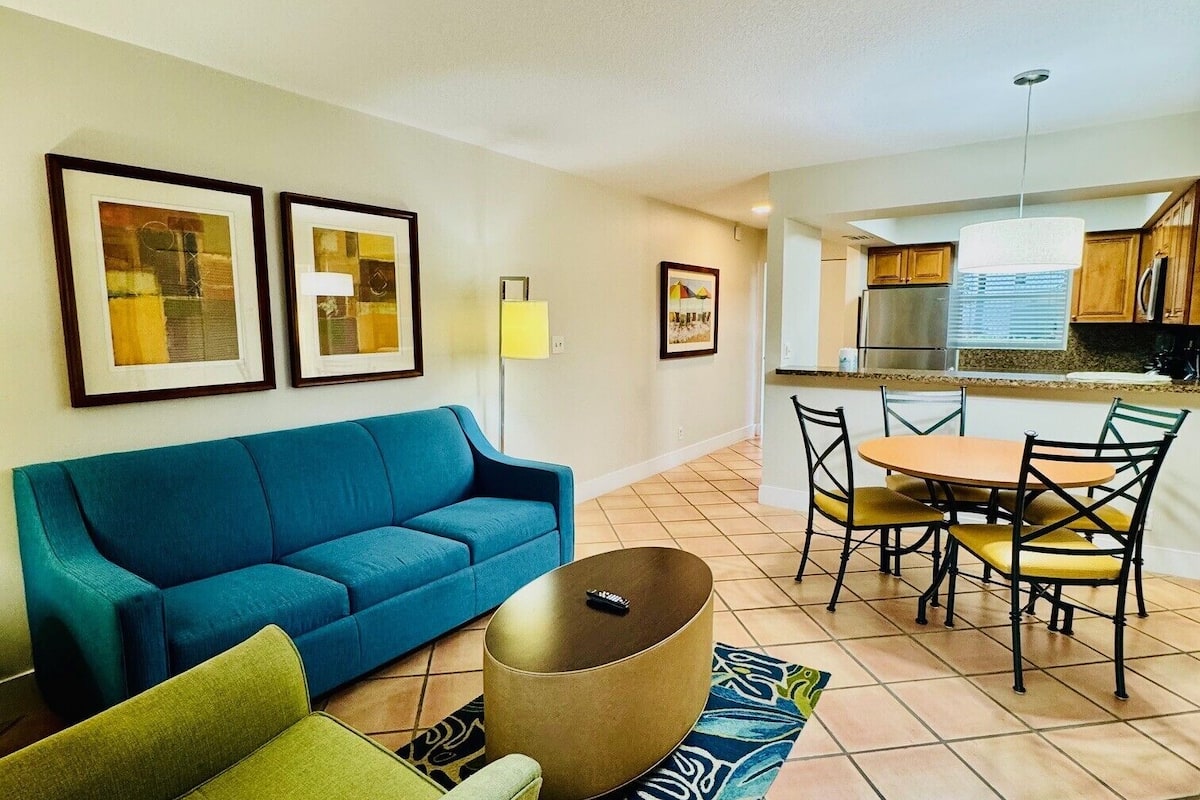 A living room with a blue sofa, a green armchair, a round coffee table, and a dining area with a table and chairs.