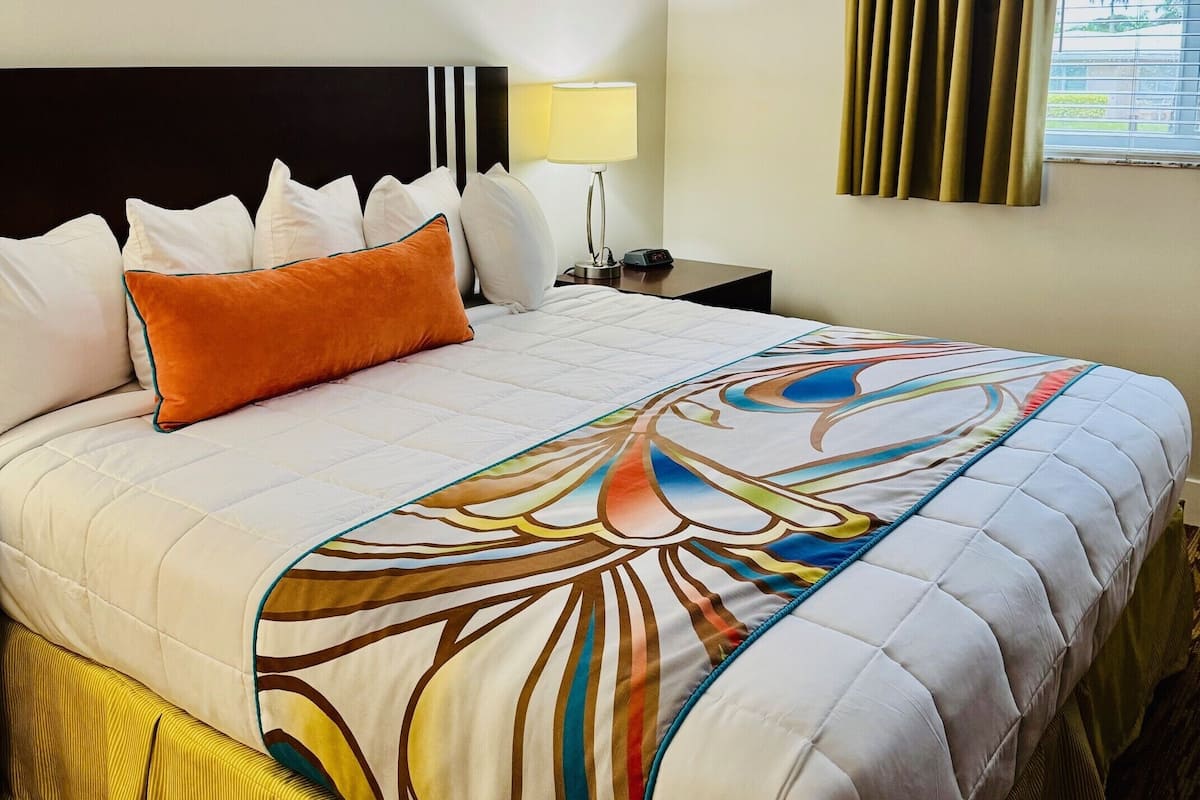 A hotel room with a large bed, a nightstand with a lamp, a window with curtains, and a view of palm trees outside.