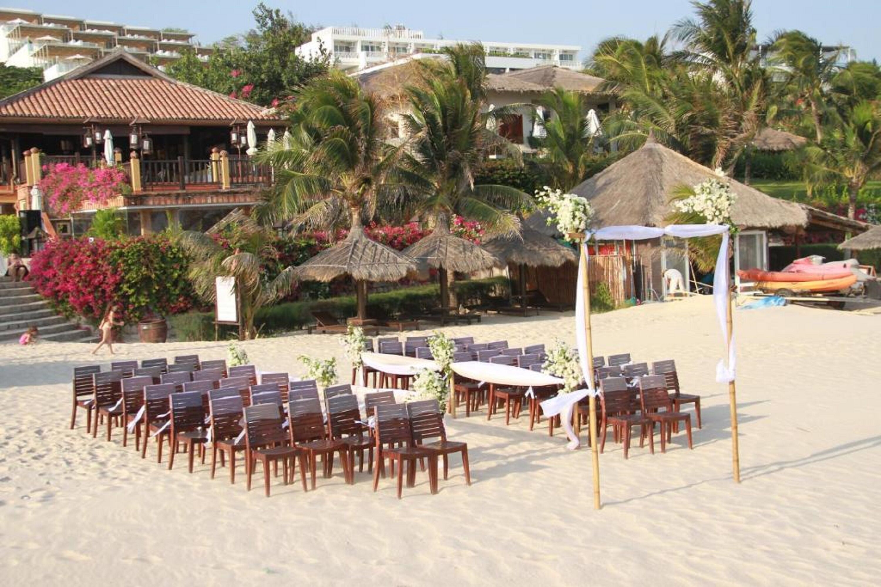 Een bruiloftdecoratie op het strand met houten stoelen en tafels, een rieten constructie en palmbomen.