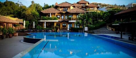 Una piscina con superficie de agua azul cristalina, rodeada de edificios con tejados de azulejos y balcones, y abundante vegetación al fondo.