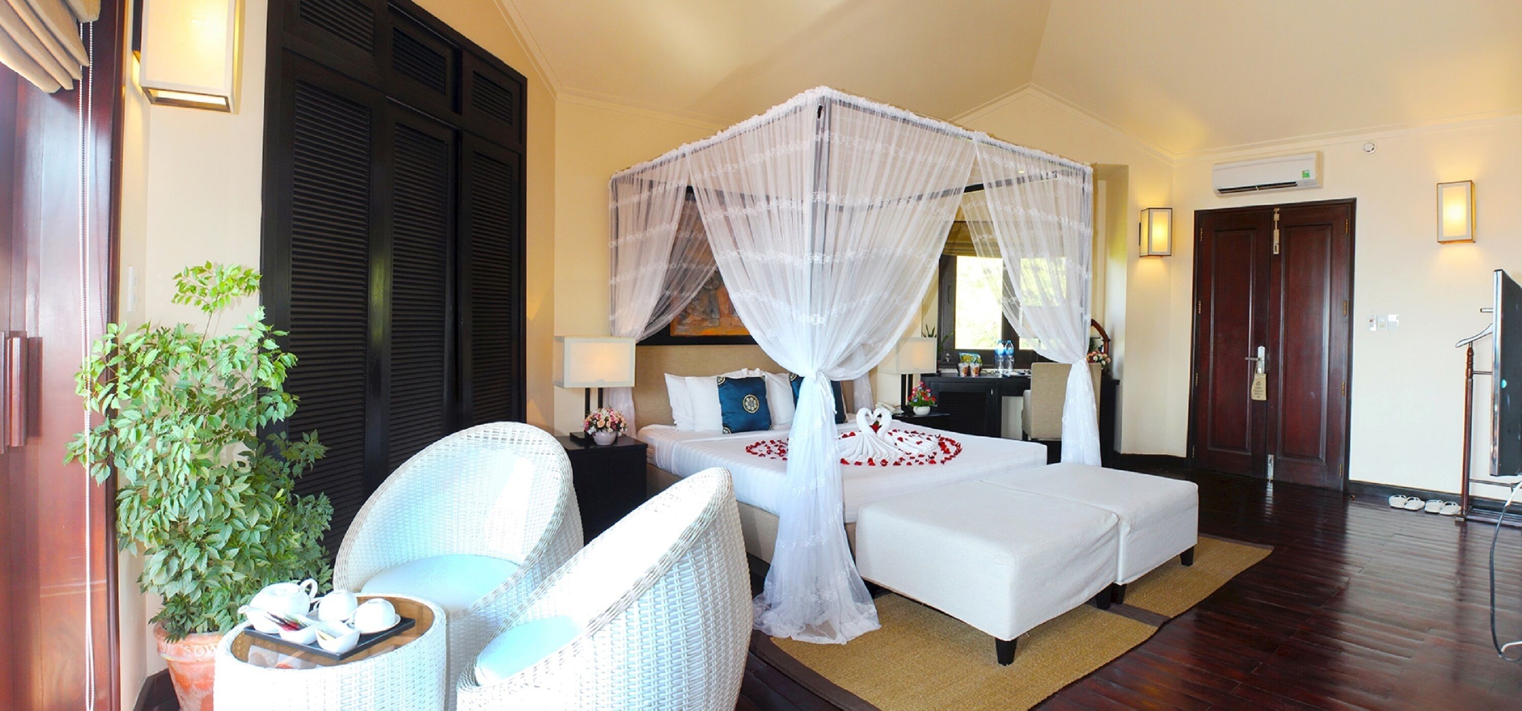 Une chambre d’hôtel avec un lit à baldaquin, un coin salon comprenant un fauteuil et une petite table, une plante en pot et un téléviseur.