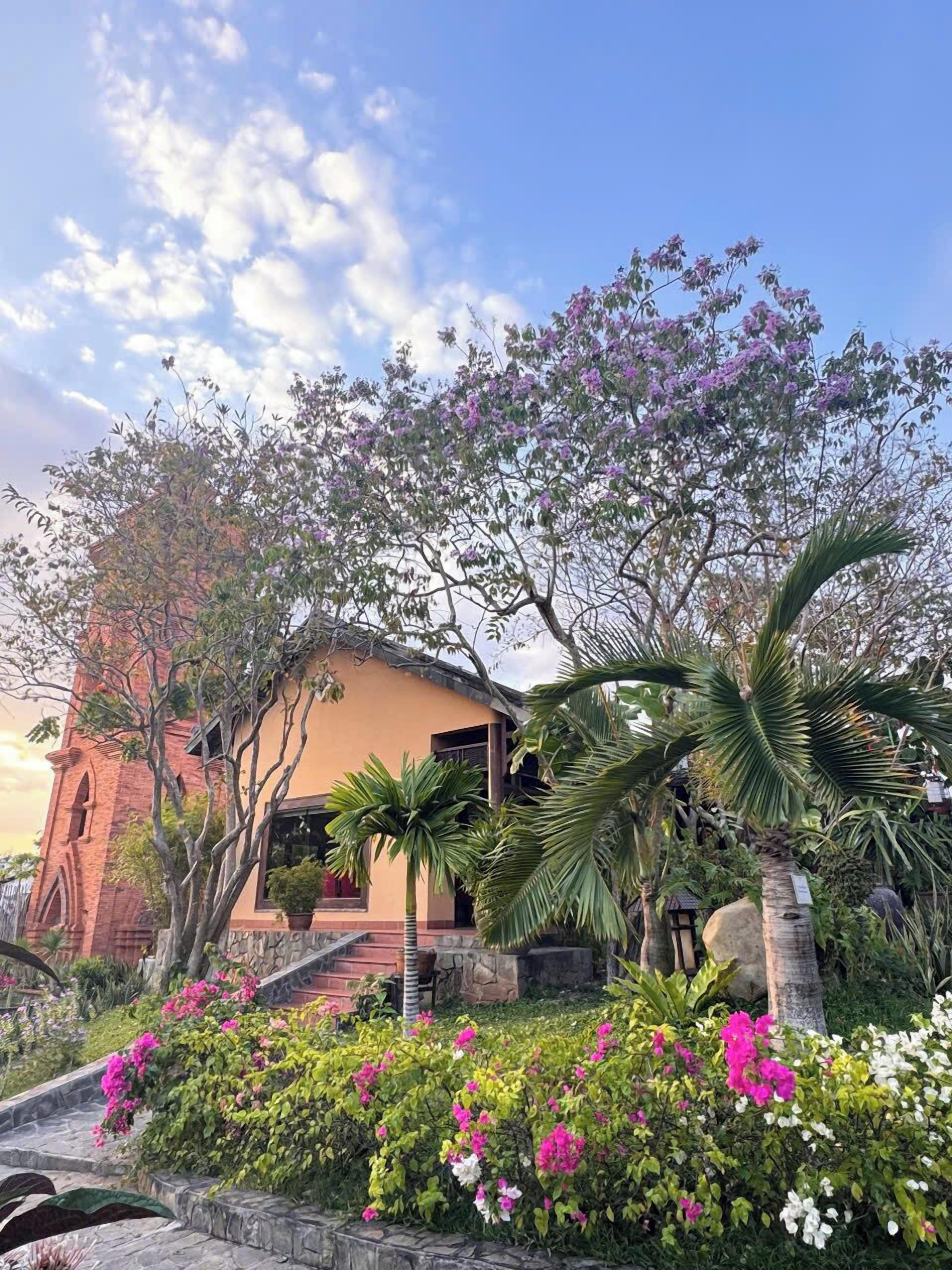 A building with a brick tower, a yellow facade, and a garden with various plants and flowers.