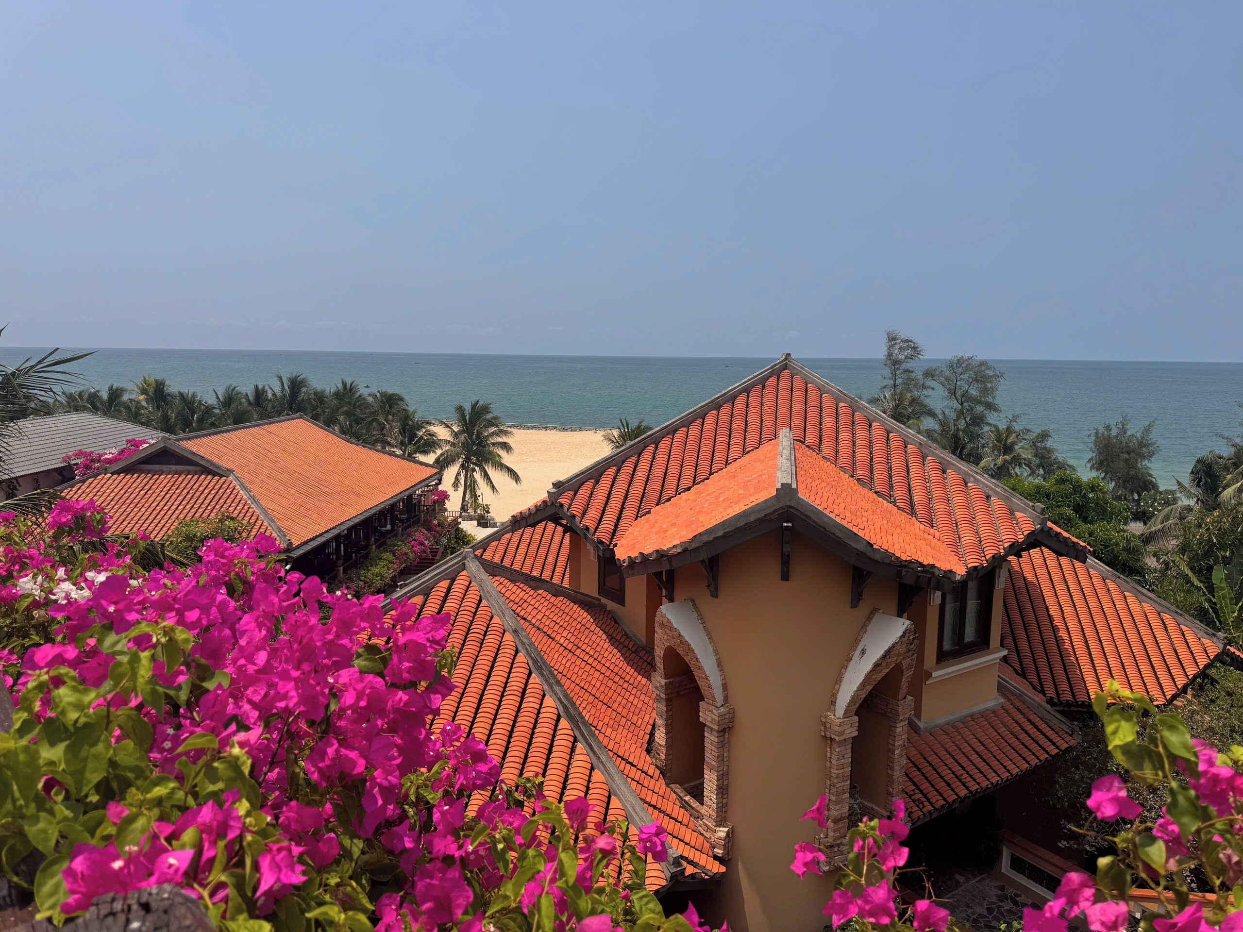 A coastal hotel with terracotta roofs, a beach view, and vibrant pink bougainvillea flowers.