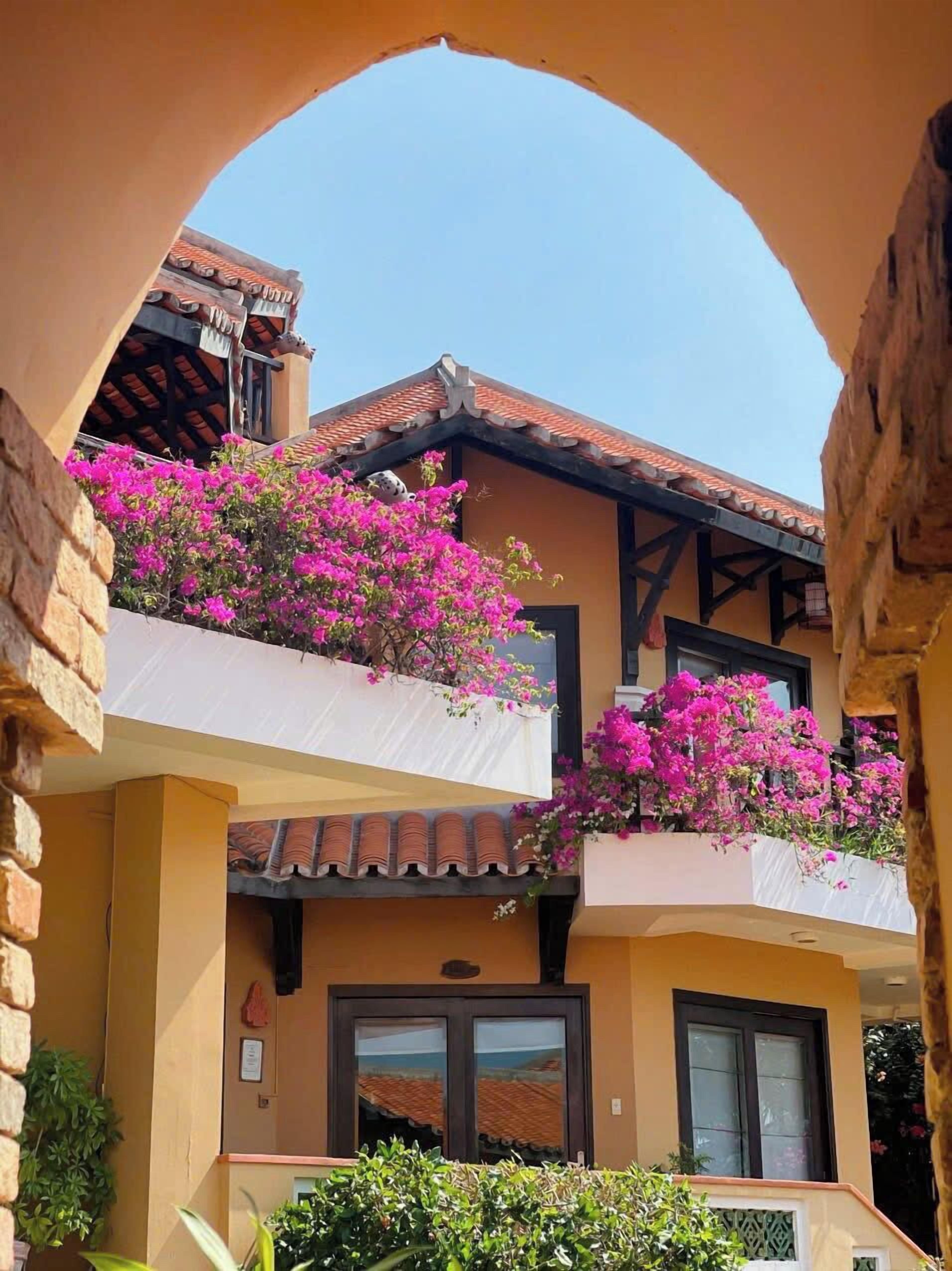 Un edificio con un balcón decorado con flores rosadas, tejado de baldosas y un cielo azul despejado.