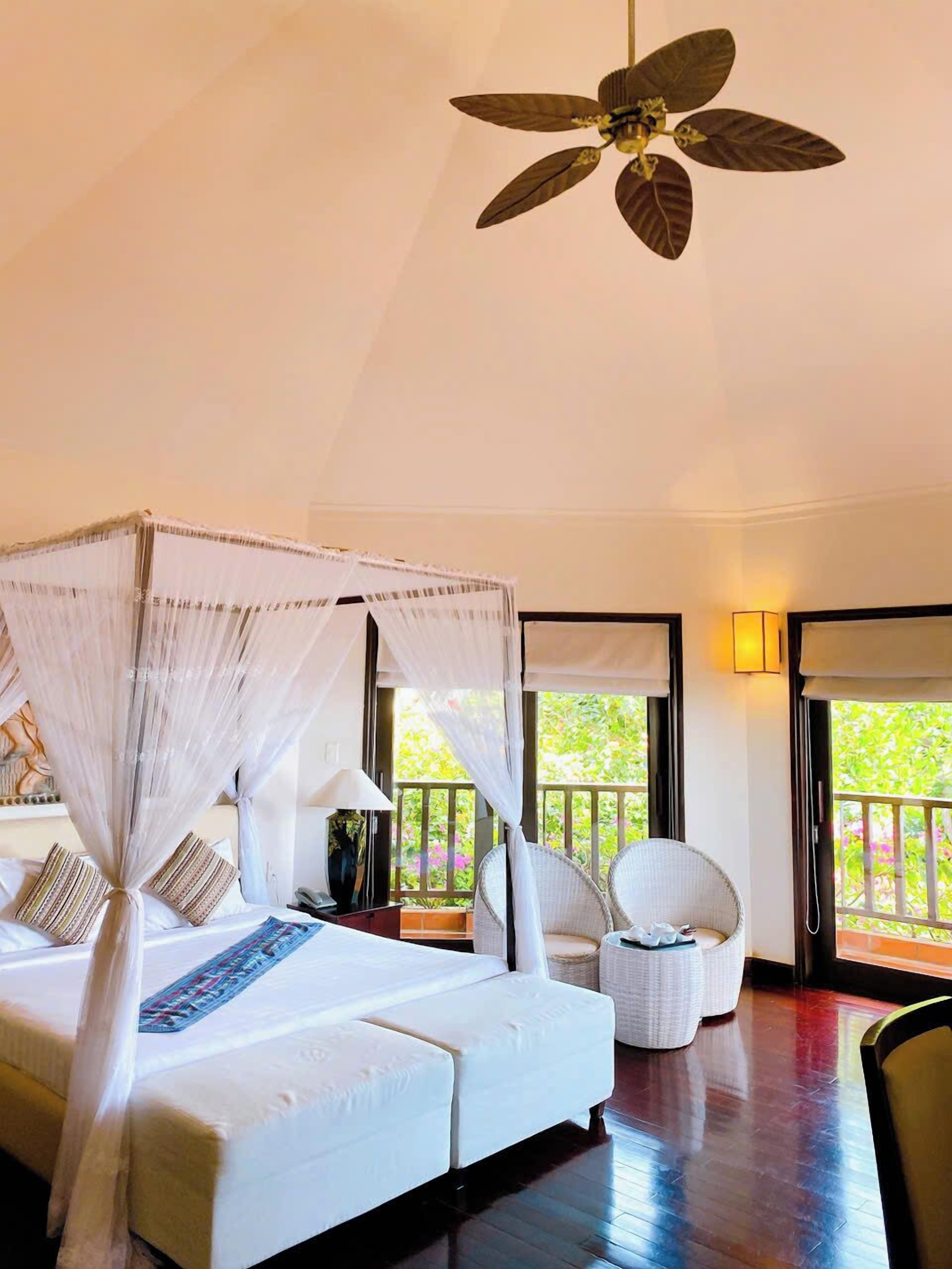 A bedroom with a four-poster bed, a ceiling fan, and a view of greenery outside.