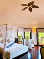 A bedroom with a four-poster bed, a ceiling fan, and a view of greenery outside.