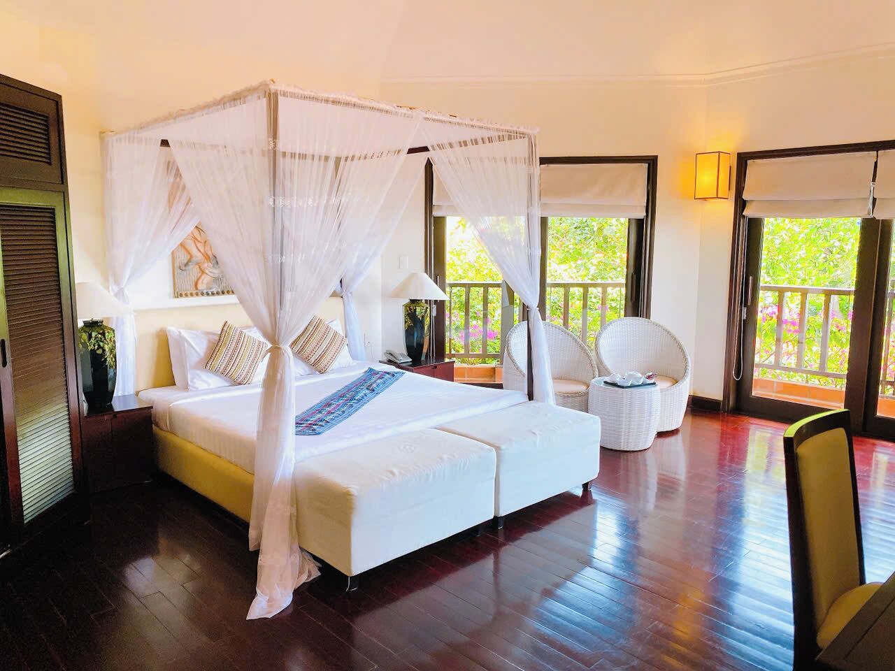 A spacious bedroom with a large bed, a sitting area with a chair, and a view of greenery outside.