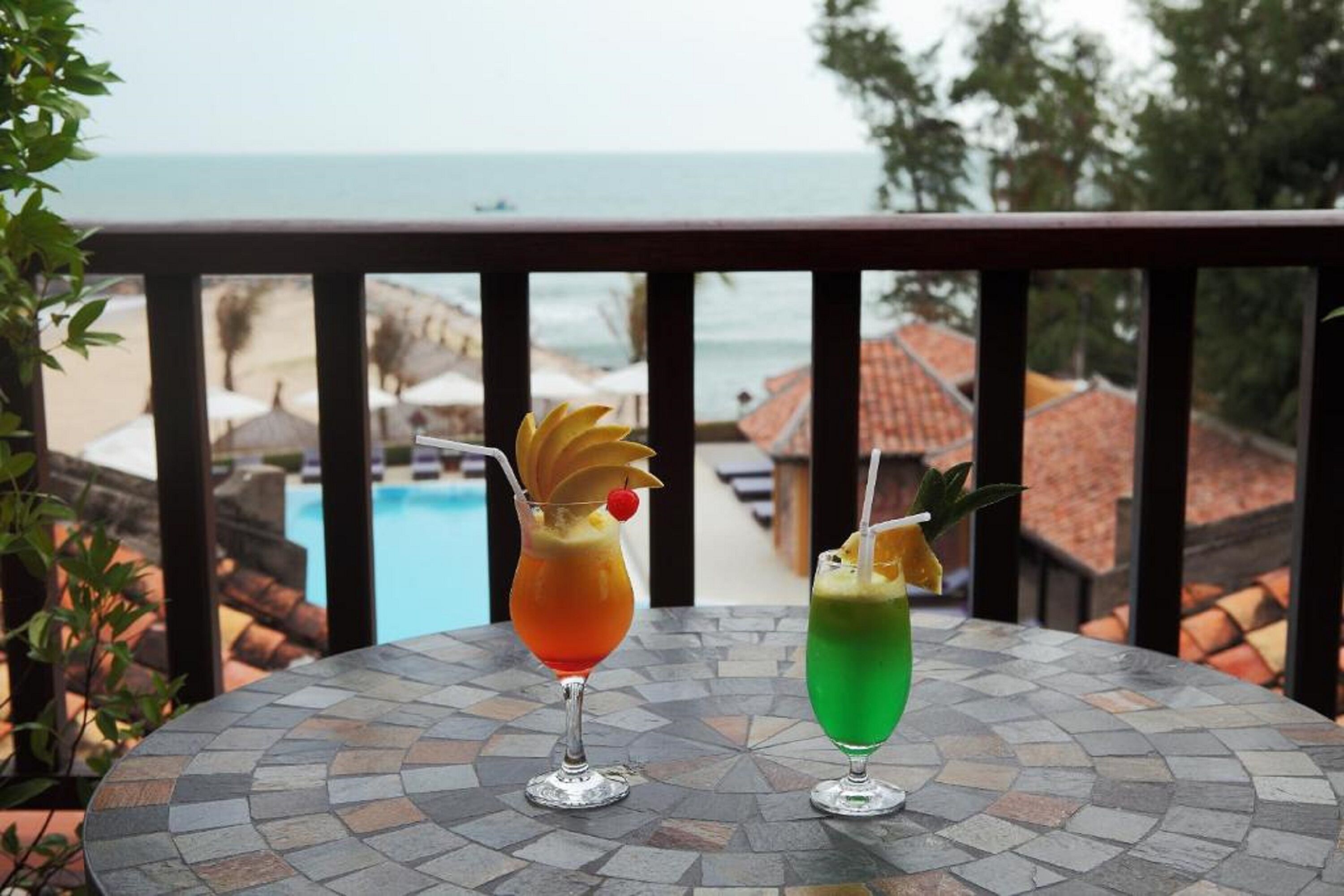 Two cocktails on a mosaic table with a view of a pool and beach in the background.