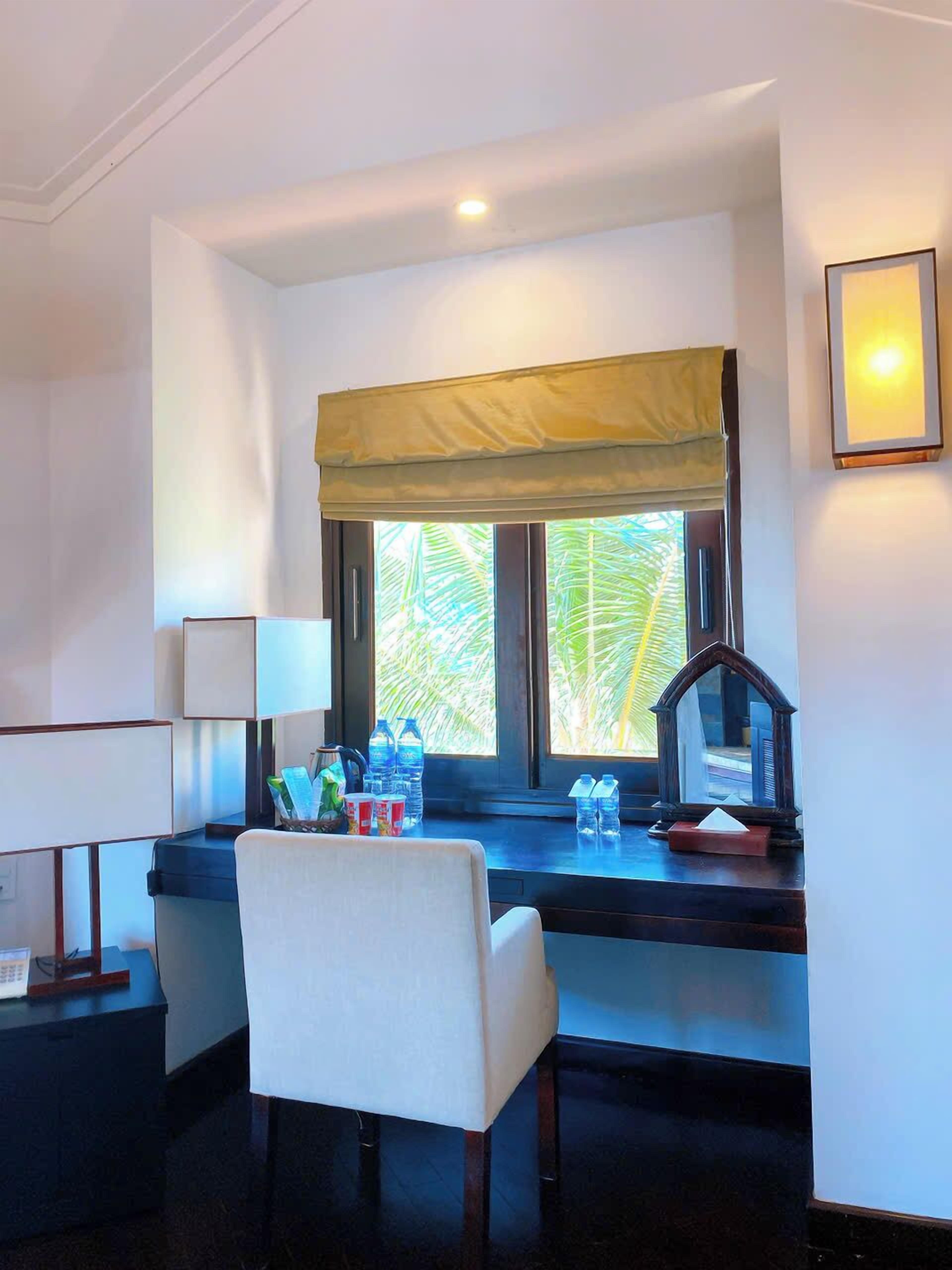 A hotel room with a desk, chair, window with a view of palm trees, and a lamp.
