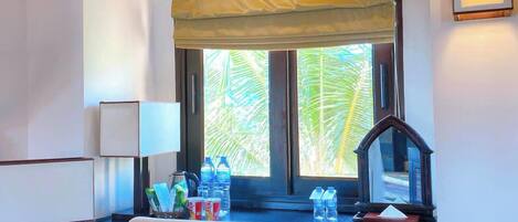 A hotel room with a desk, chair, window with a view of palm trees, and a lamp.
