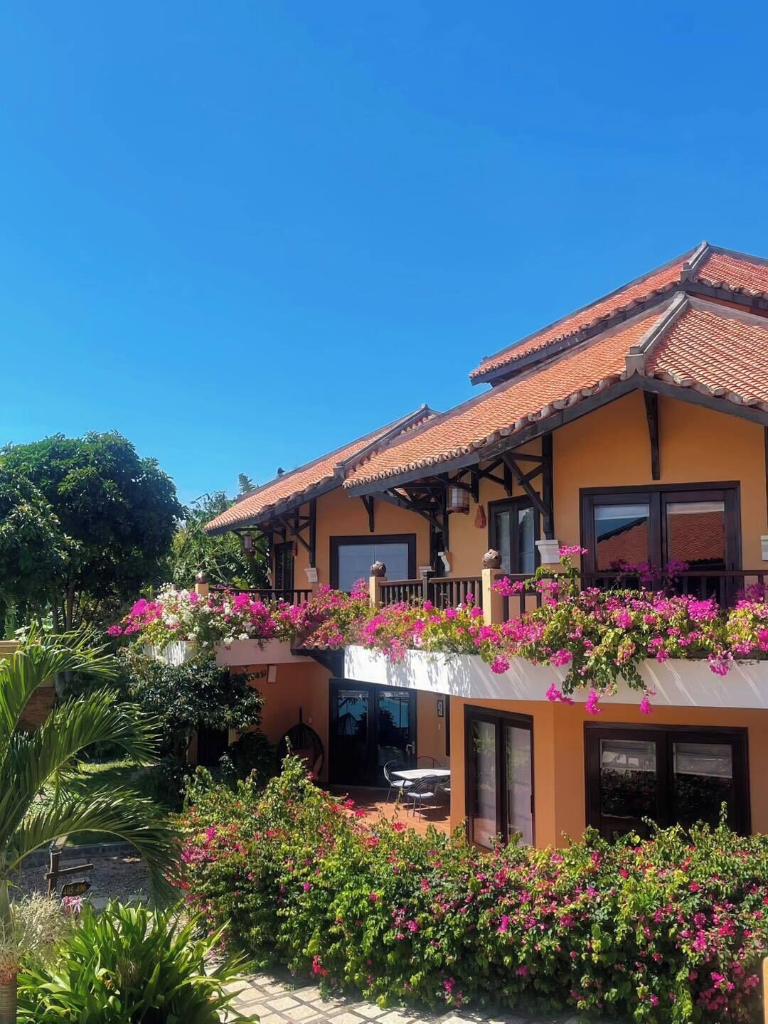 A two-story building with a tiled roof, balconies with flower boxes, and a garden area with various plants.