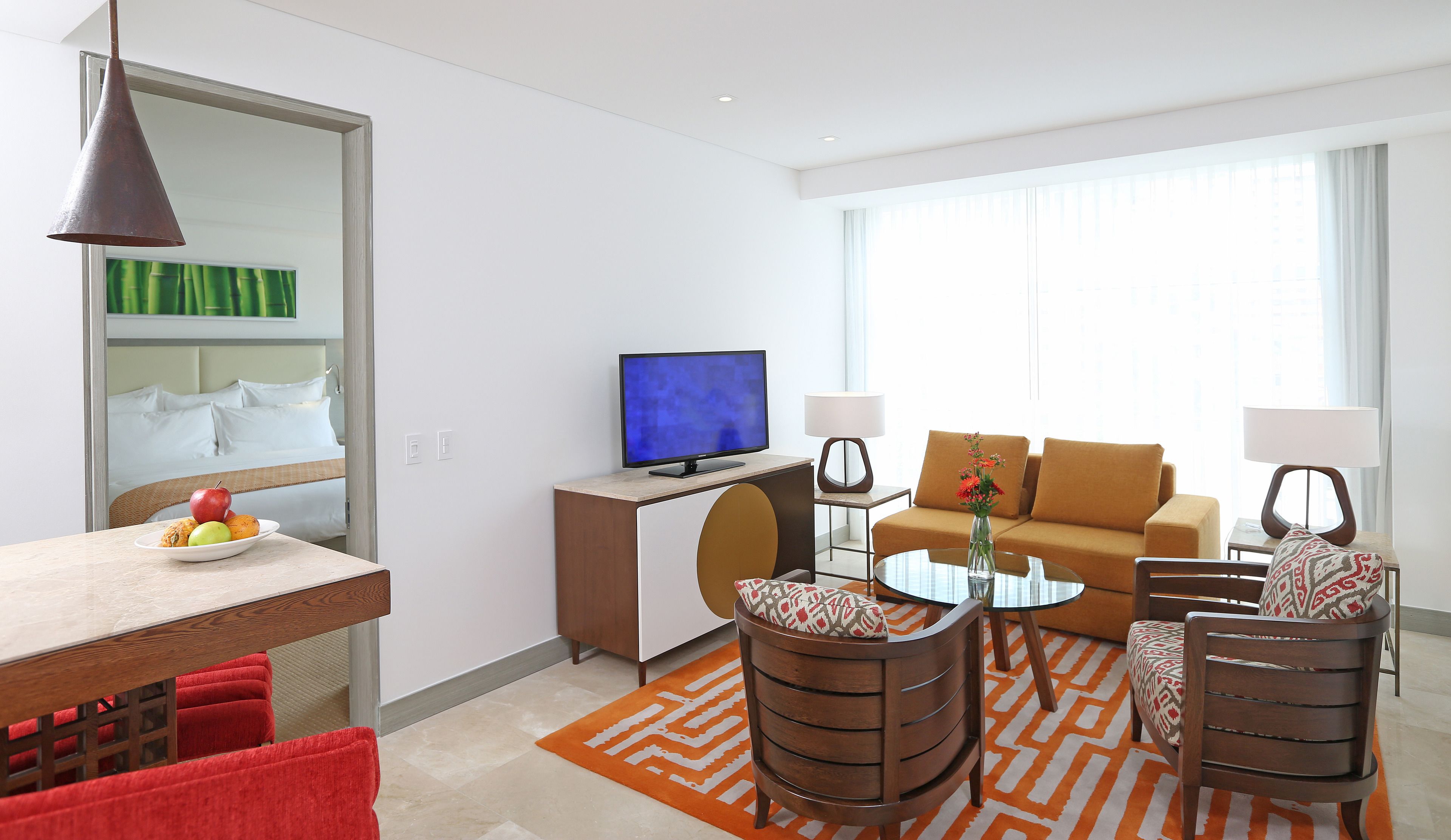 A modern hotel room with a bed, a TV, a sofa, a coffee table, and a dining area with a bowl of fruit.
