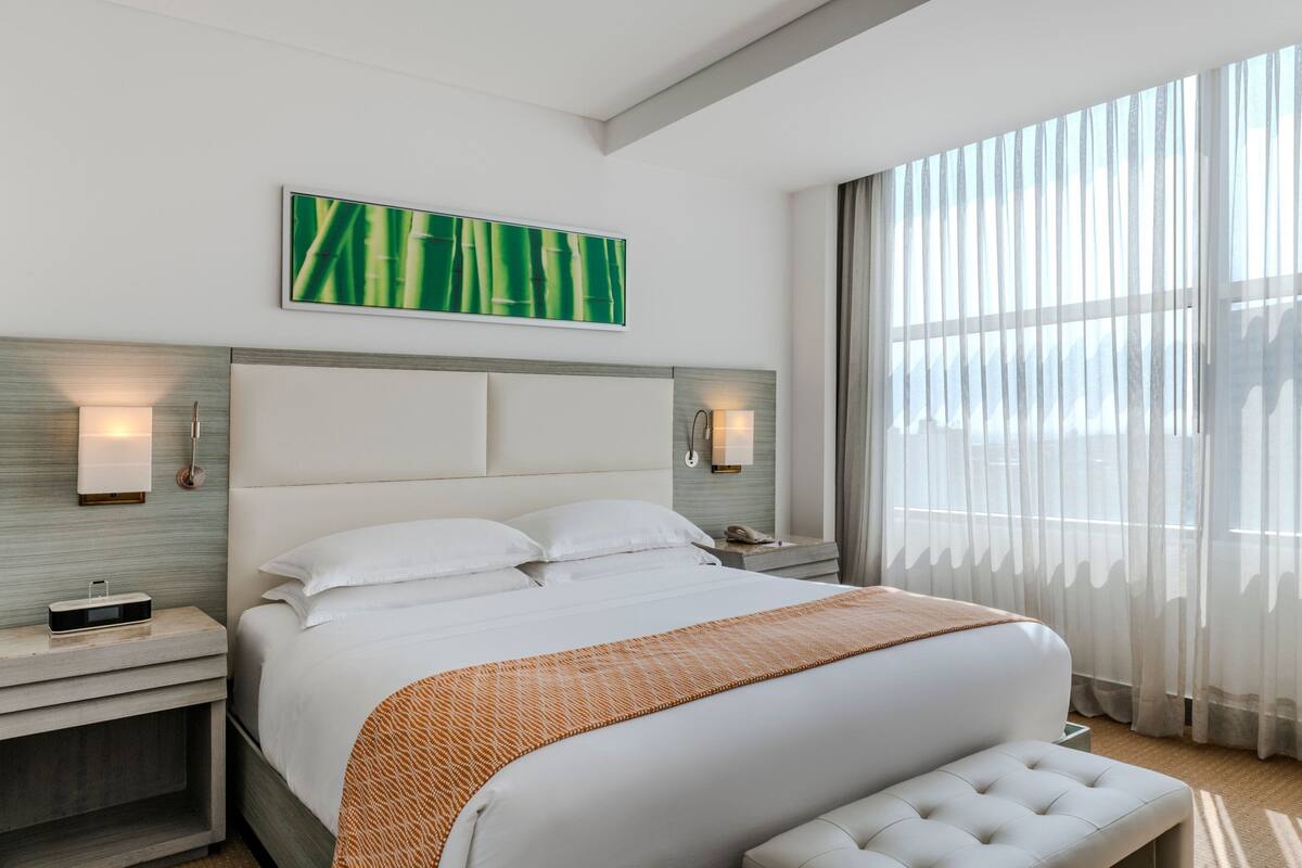 Ein modernes Hotelzimmer mit einem großen Bett, einer Kopfteilbeleuchtung, einem Nachttisch mit Telefon und Blick ins Freie durch transparente Vorhänge.