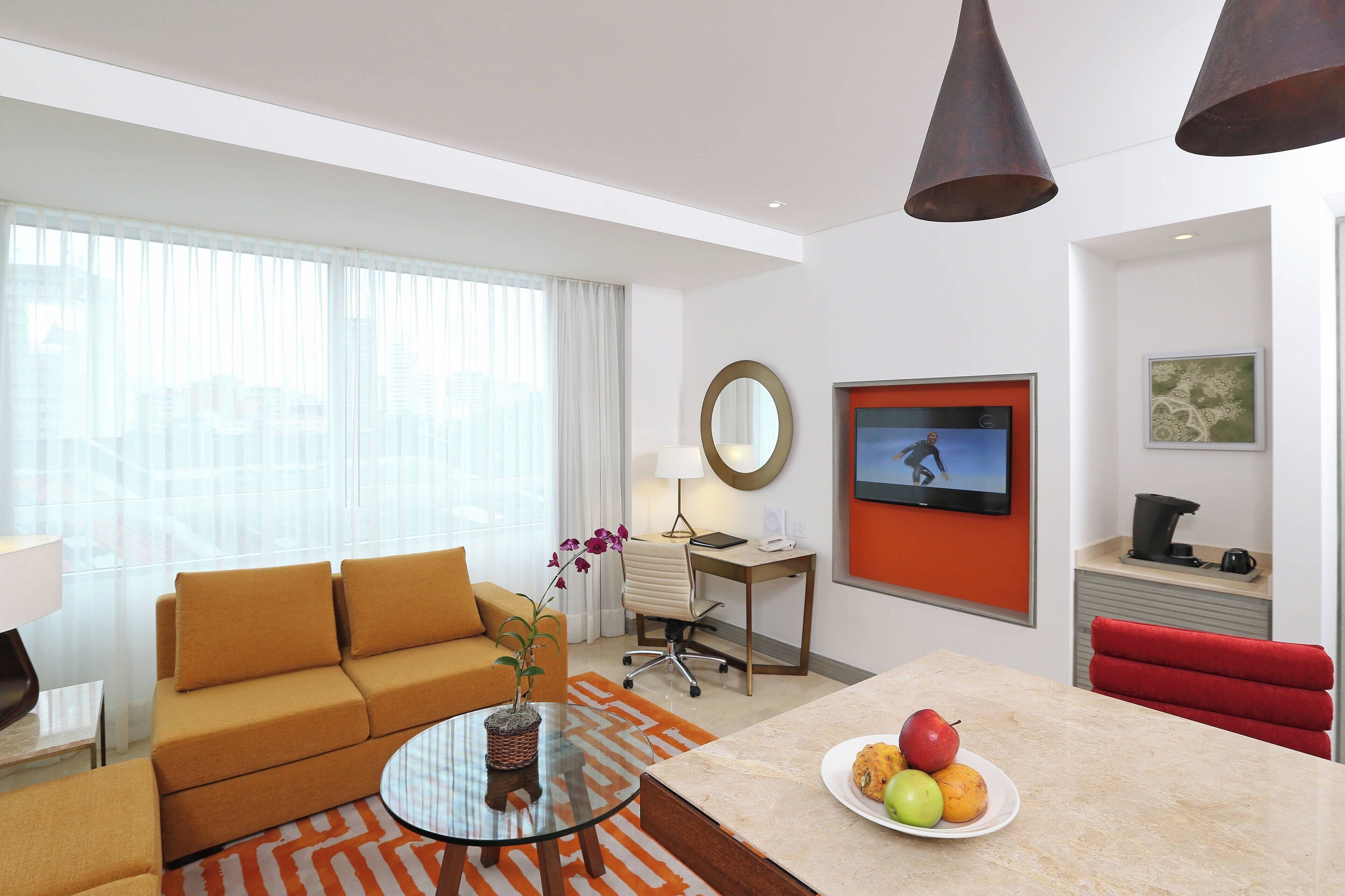 A modern hotel room with a sofa, a round glass table with a plant, a desk with a chair, a television, and a red chair.