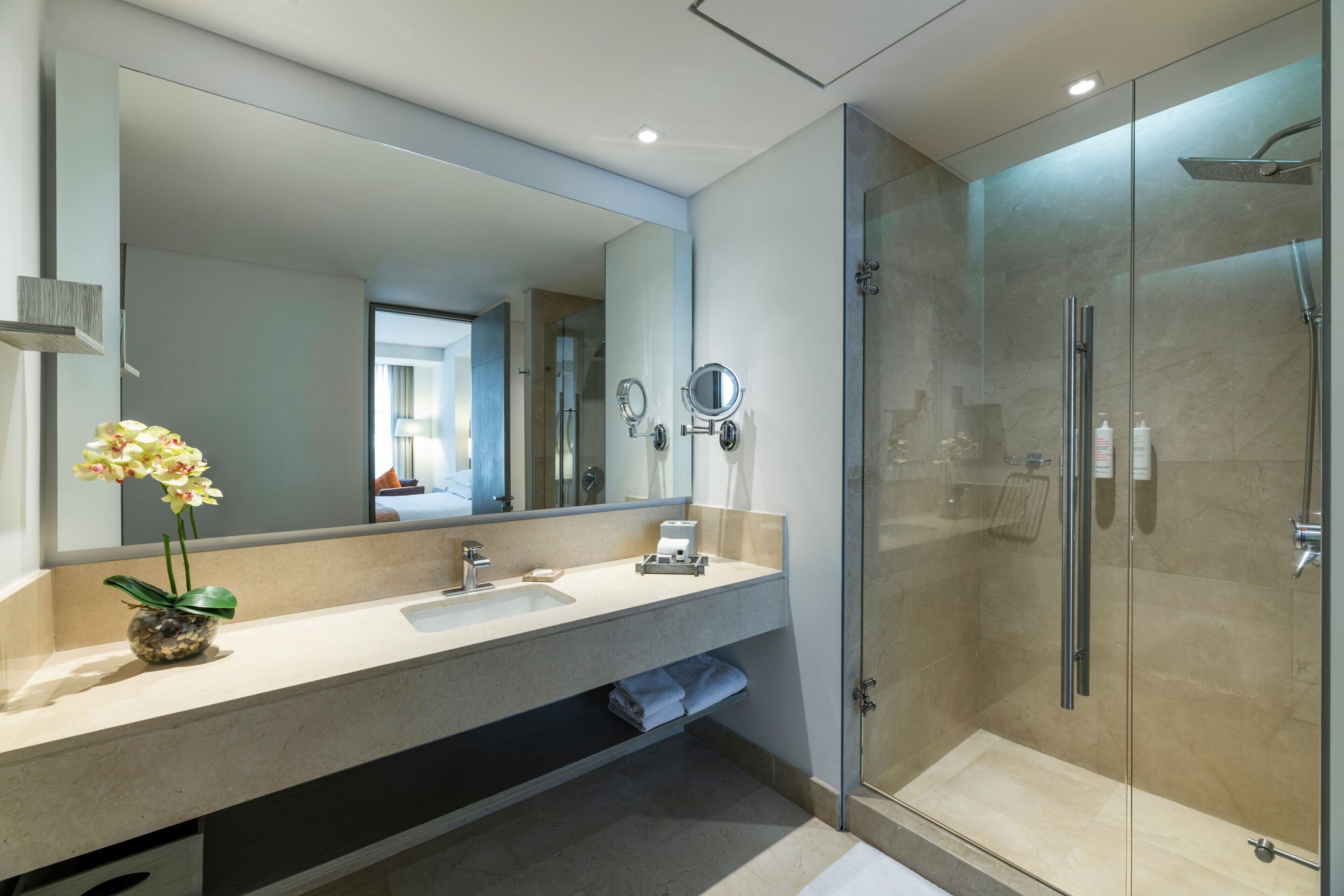 Un baño moderno con un espejo grande, un lavabo, una ducha y un florero con flores.