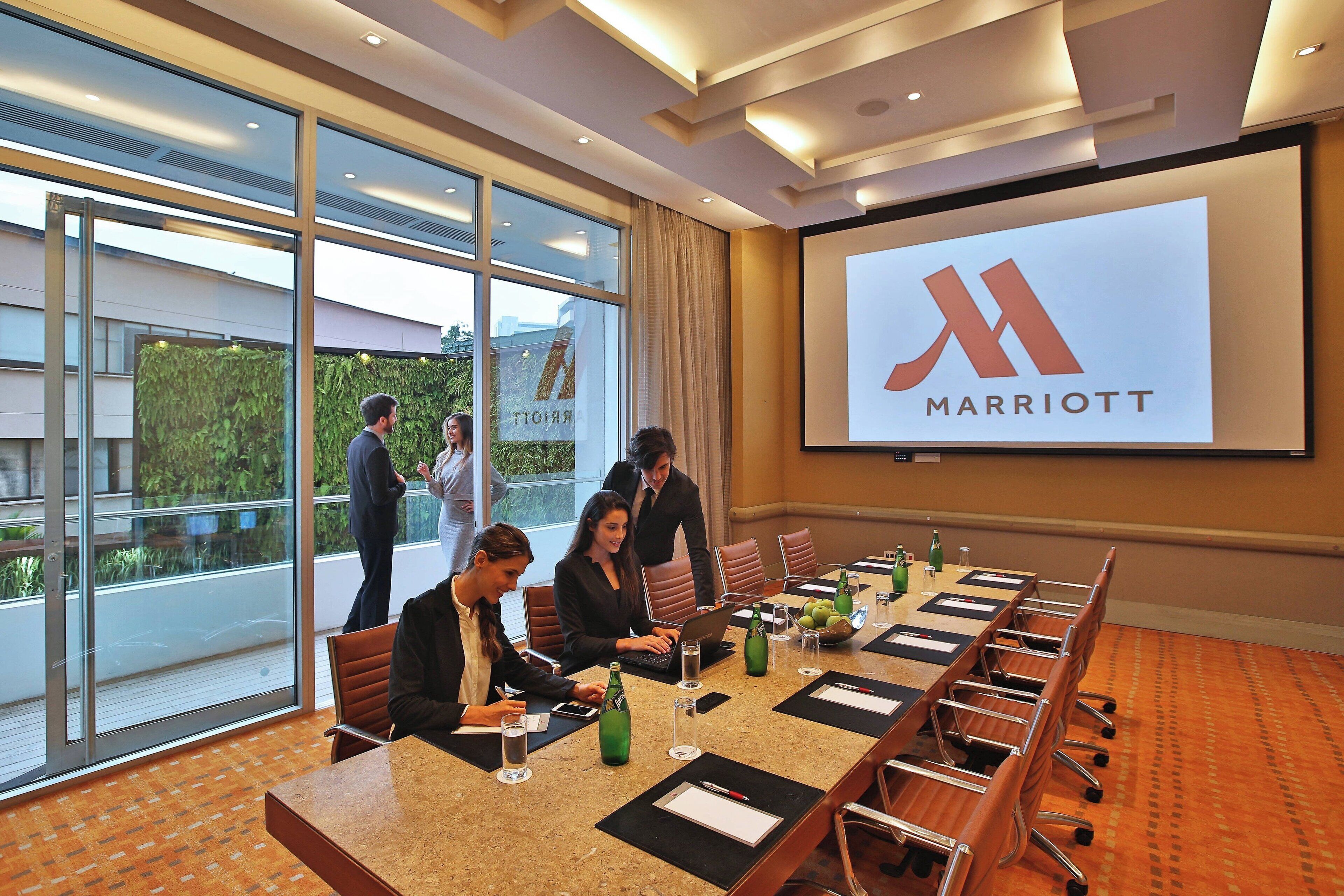 A conference room with a large screen displaying the Marriott logo, a long table with chairs, and a glass wall offering a view of the outdoors.
