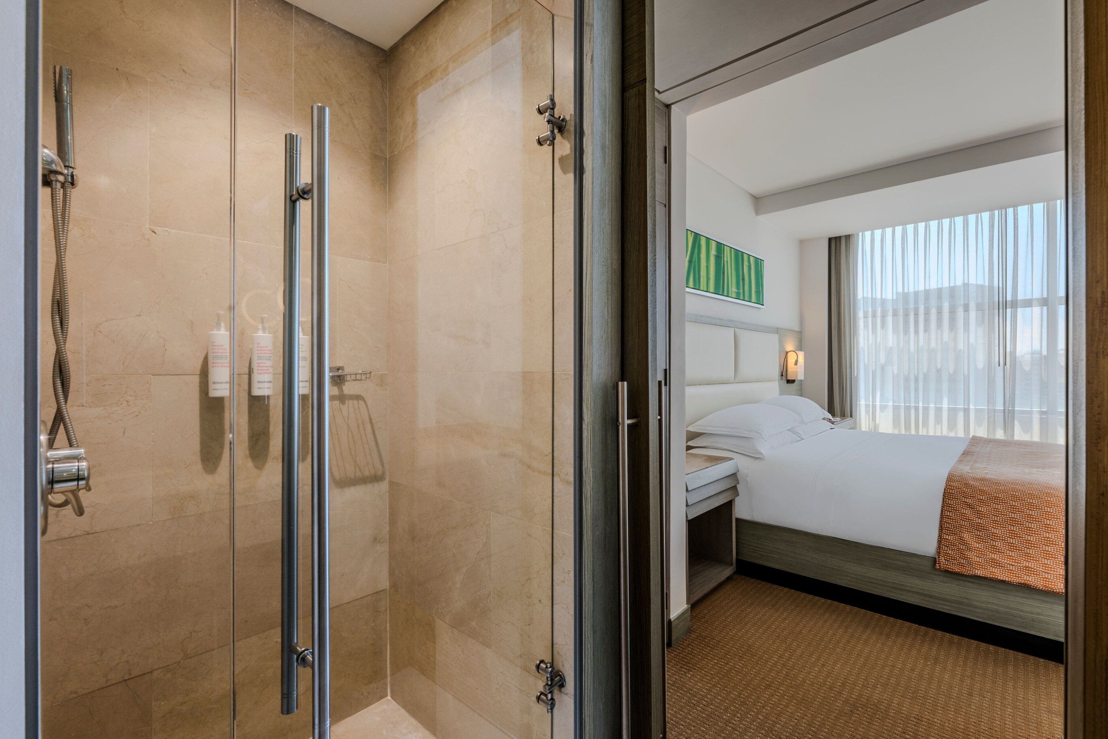 Une chambre d’hôtel dotée d’une douche à parois de verre, d’un lit avec une tête de lit blanche et offrant une vue sur la ville grâce à de grandes fenêtres.