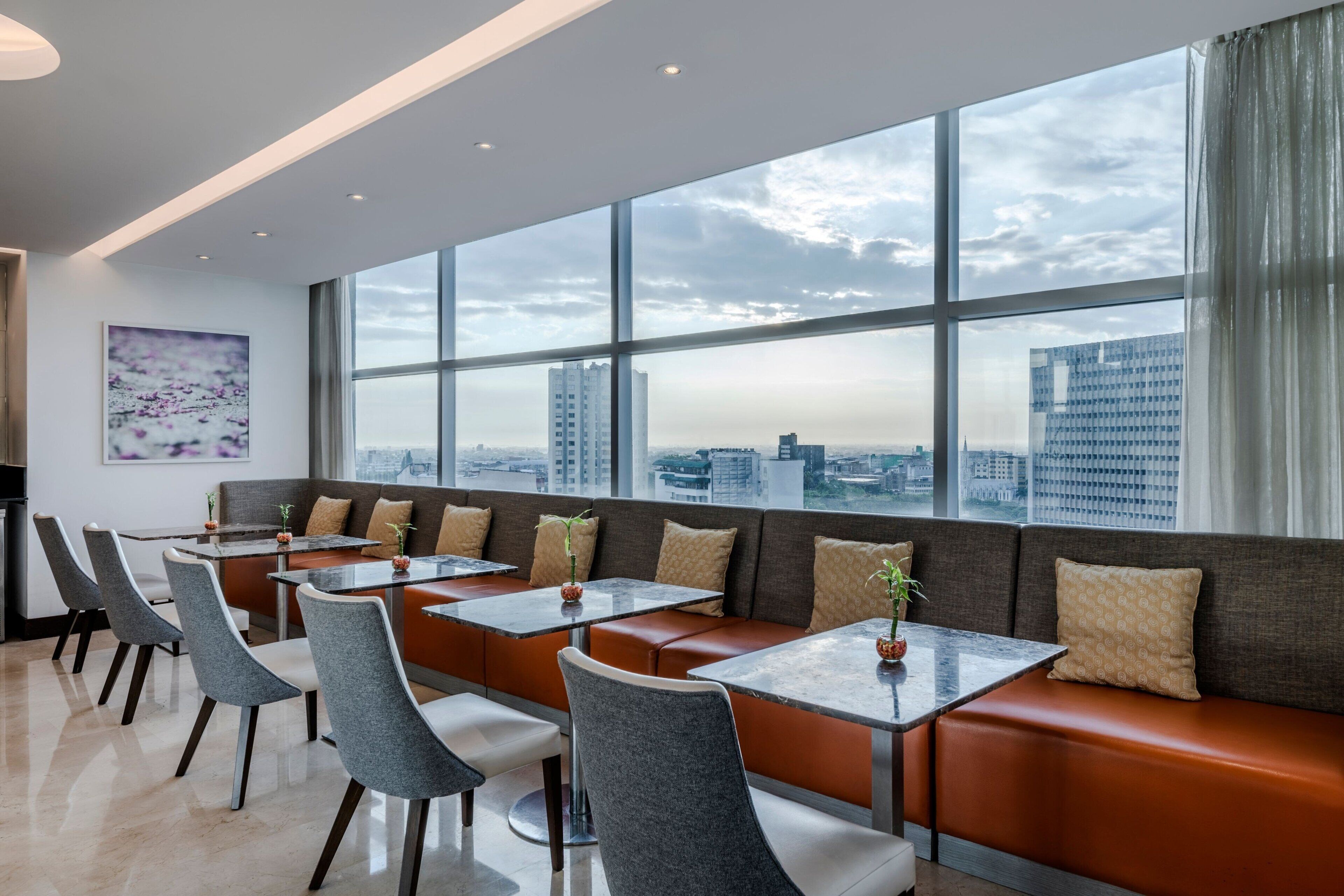 A modern dining area with a city view, featuring a long bench with cushioned seating and individual tables.