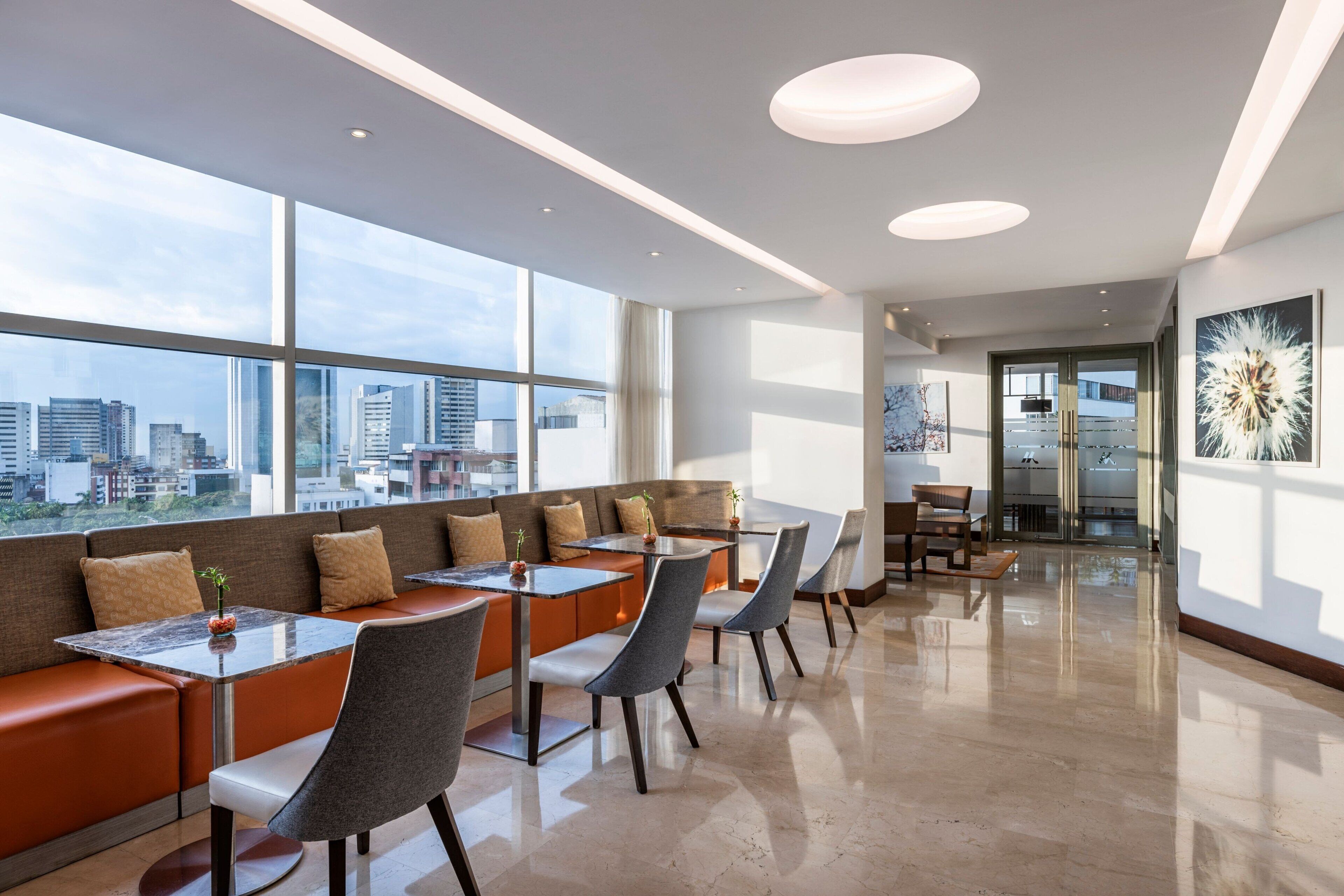 A modern dining area with high ceilings, large windows, and a city view.