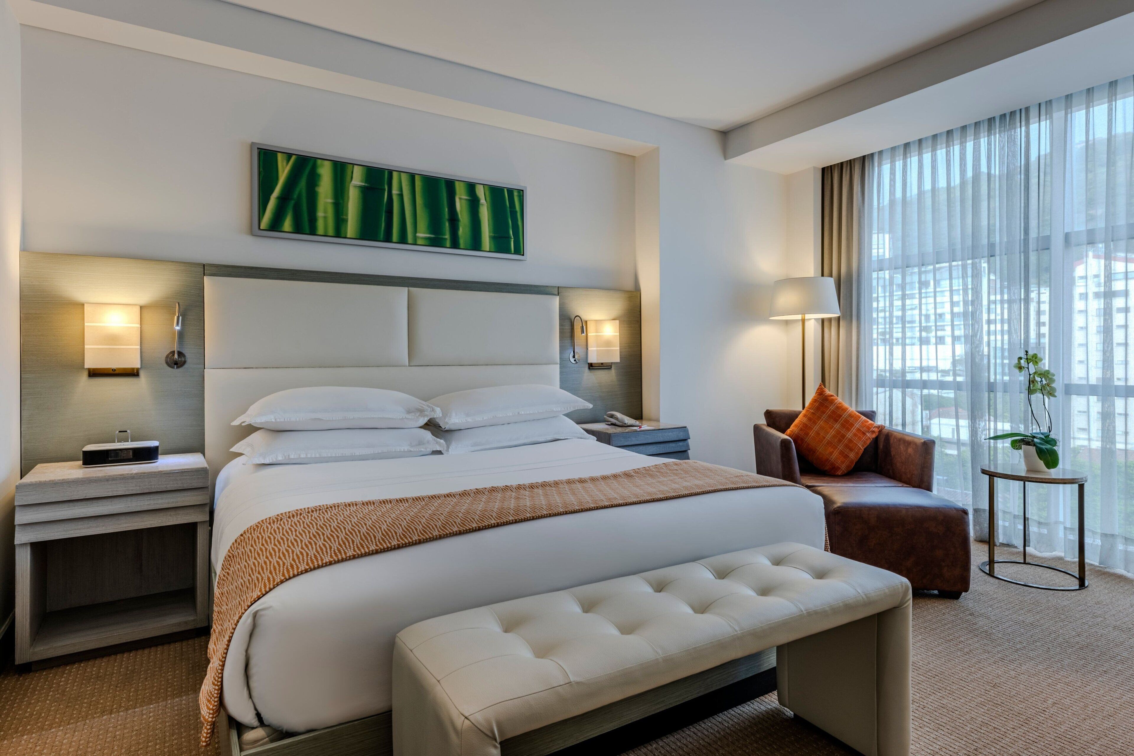 A modern hotel room with a large bed, a bench, a sofa, and a small table with a plant.