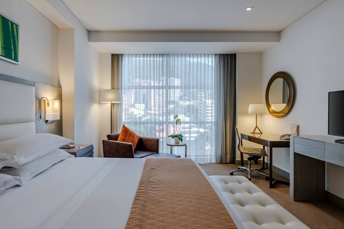 Ein Hotelzimmer mit einem großen Bett, einem Schreibtisch, einem Sessel, einem Fernseher, einer Lampe und Blick auf die Stadt durch das Fenster.