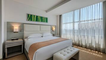 Une chambre d’hôtel moderne avec un grand lit, une banquette, une table de chevet et une vue sur la ville à travers des rideaux transparents.