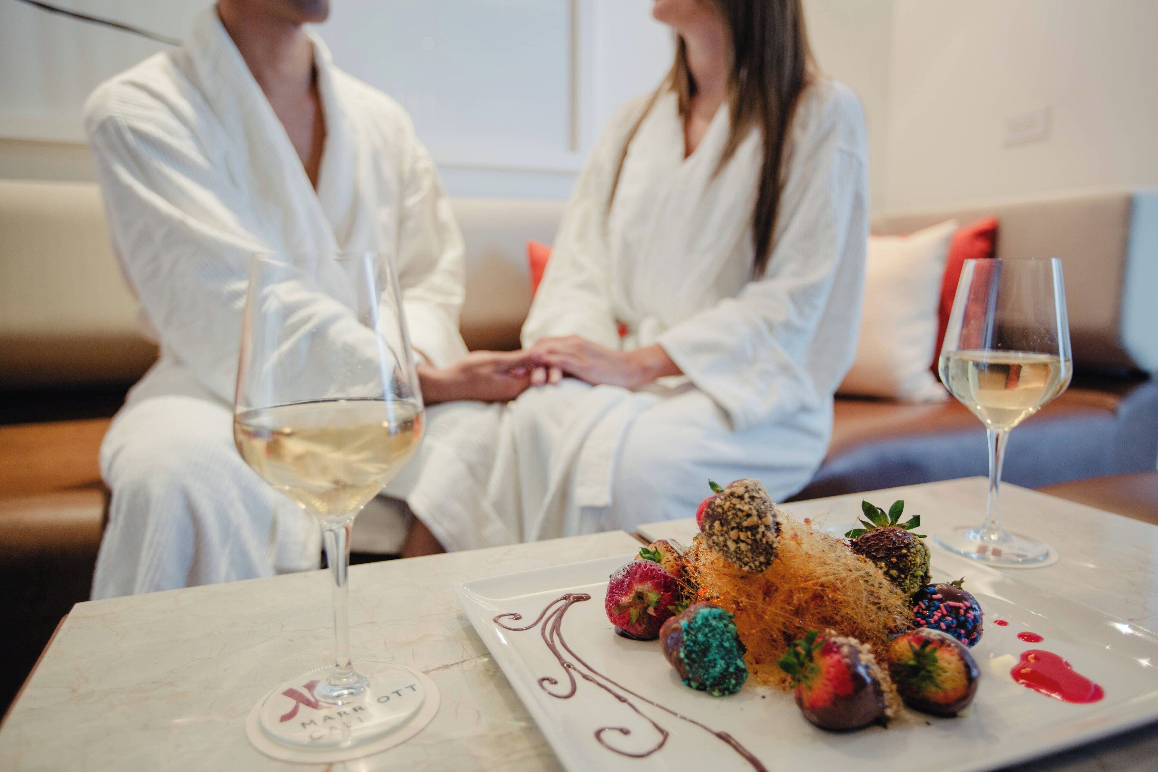 Una pareja vestida de blanco sentada en un sofá, con un plato de postre y copas de vino sobre una mesa.