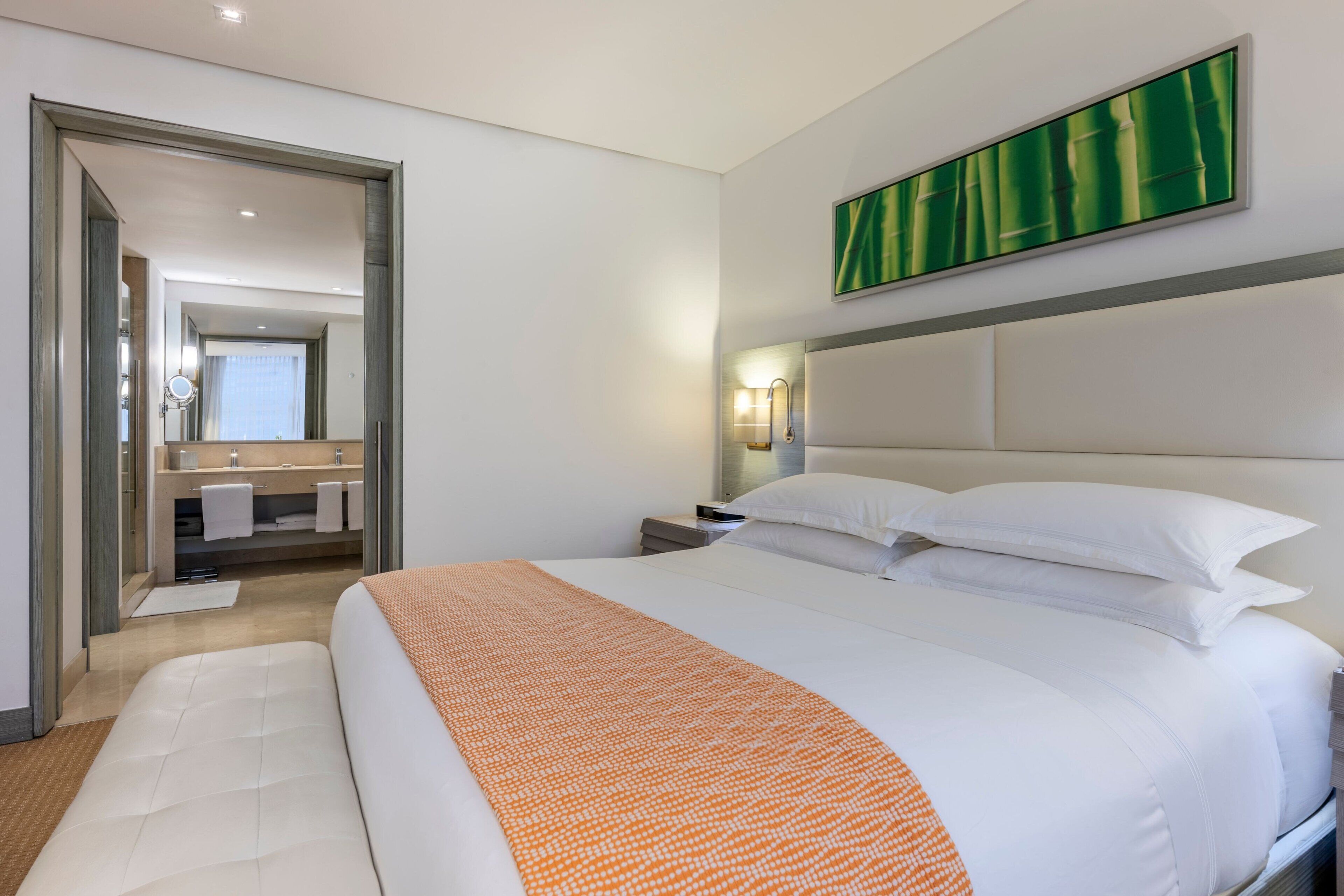Une chambre d’hôtel moderne avec un grand lit, une salle de bain dotée d’un miroir et d’un porte-serviettes, et un tableau abstrait aux couleurs vertes accroché au mur.