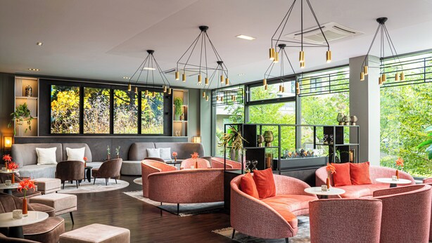 Een moderne zithoek met banken en fauteuils, grote ramen met uitzicht op groen, en hanglampen.