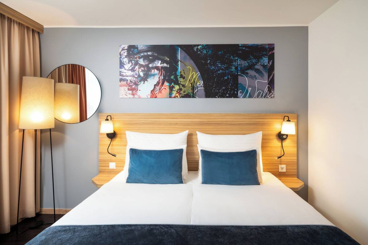A hotel room with a large bed, two bedside lamps, a round mirror, and a colorful abstract painting on the wall.