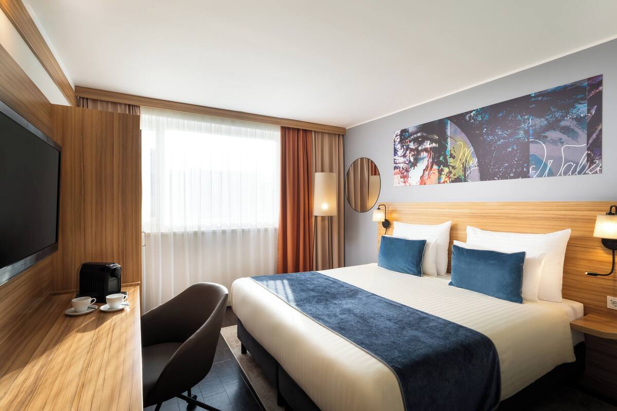 A hotel room with a large bed, a desk with a coffee maker, a TV, and a wall-mounted artwork.