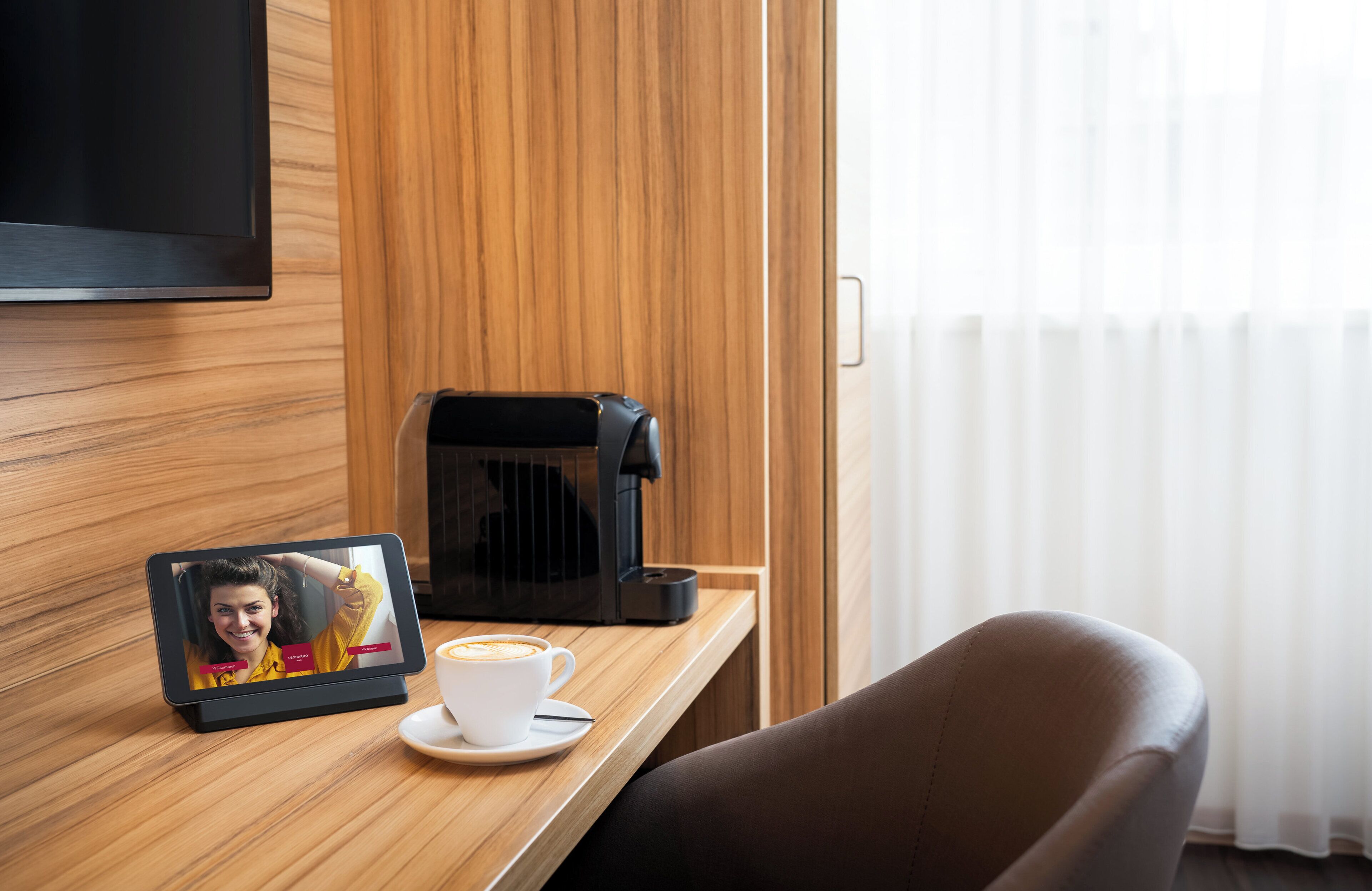 A hotel room with a wooden panel wall, a flat-screen TV mounted above a wooden desk, a coffee maker, a cup of coffee, and a tablet displaying a photo.