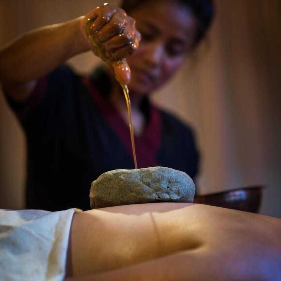 A person pouring a liquid onto a stone placed on a person's back.