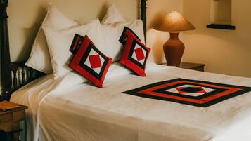 Un lit bien fait, recouvert d’une couvre-lit blanche et agrémenté de deux coussins rouges et noirs, une table de chevet en bois avec une lampe, et un tapis à motifs posé sur un sol carrelé.