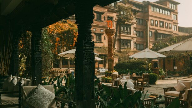 Un espacio al aire libre con techo, con sillas y mesas, rodeado de vegetación y un edificio al fondo.