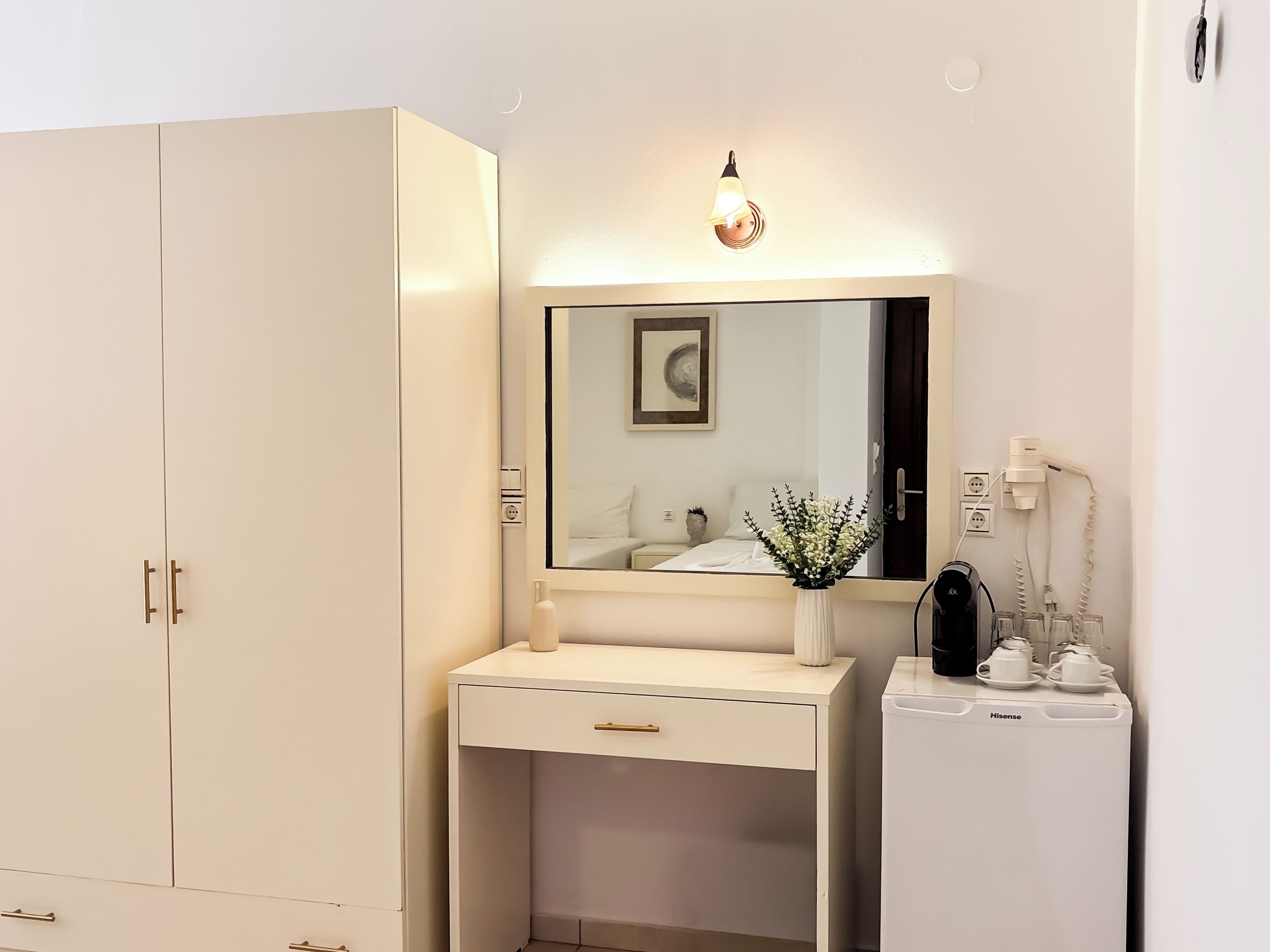 A white wardrobe with a mirror, a bedside table with a vase of flowers, and a bedside heater.