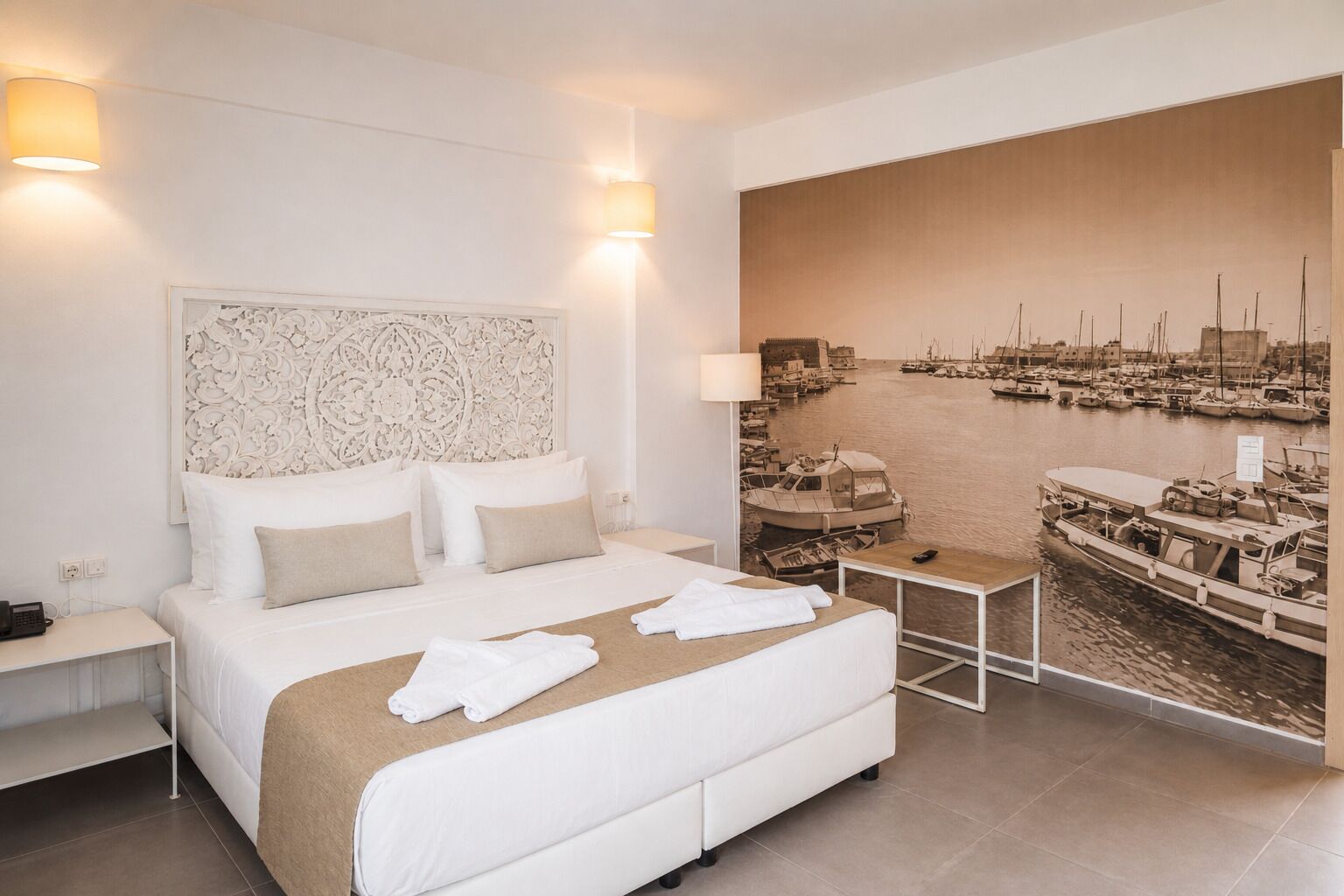 A hotel room with a bed, a nightstand, a bedside table, a wall-mounted lamp, and a large mural of a marina.