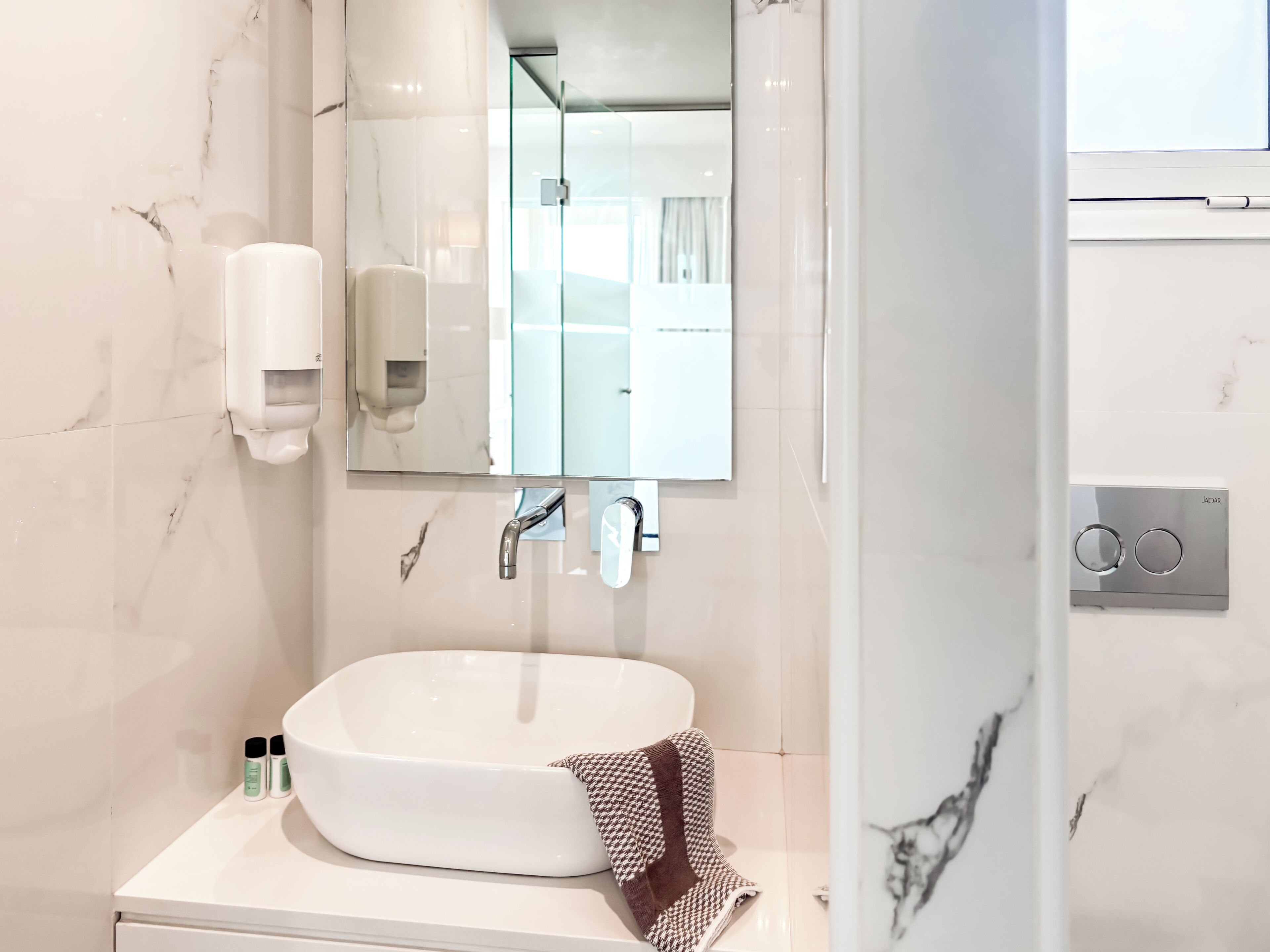 Un baño con lavamanos blanco, un espejo, una toalla y un dispensador de jabón.