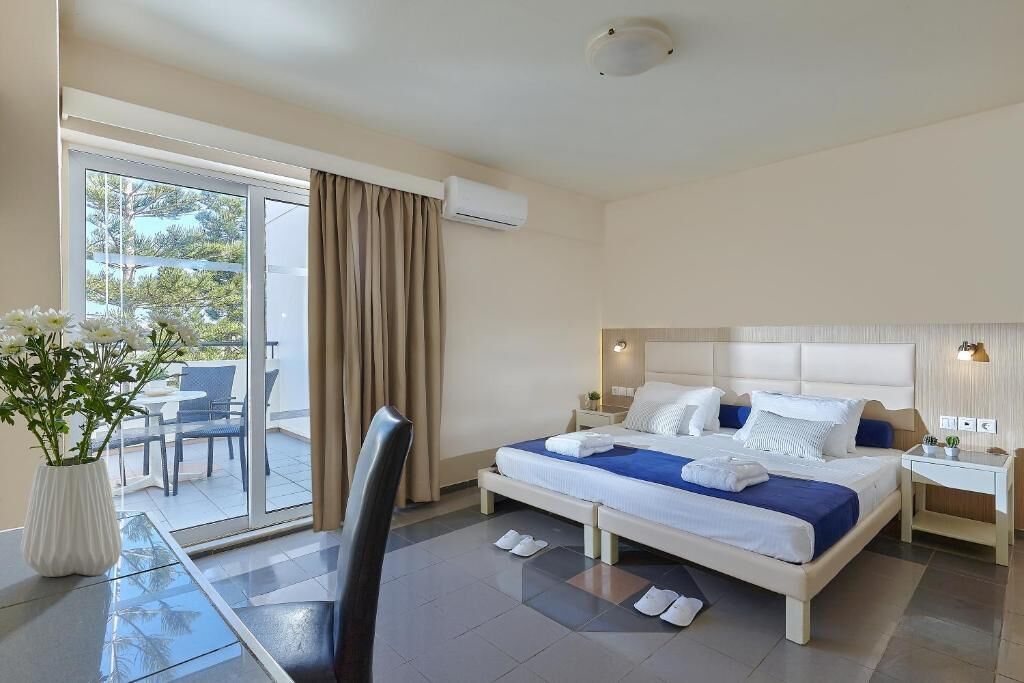 A hotel room with a bed, a chair, a table, a potted plant, and a view of the outdoors.