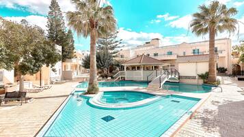 A swimming pool with a tiled deck, palm trees, and a building with a tiled roof in the background.