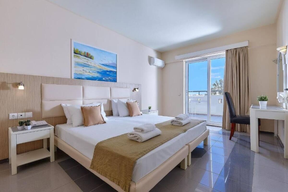 A hotel room with a bed, bedside tables, a desk, a chair, a painting, and a view through a window.