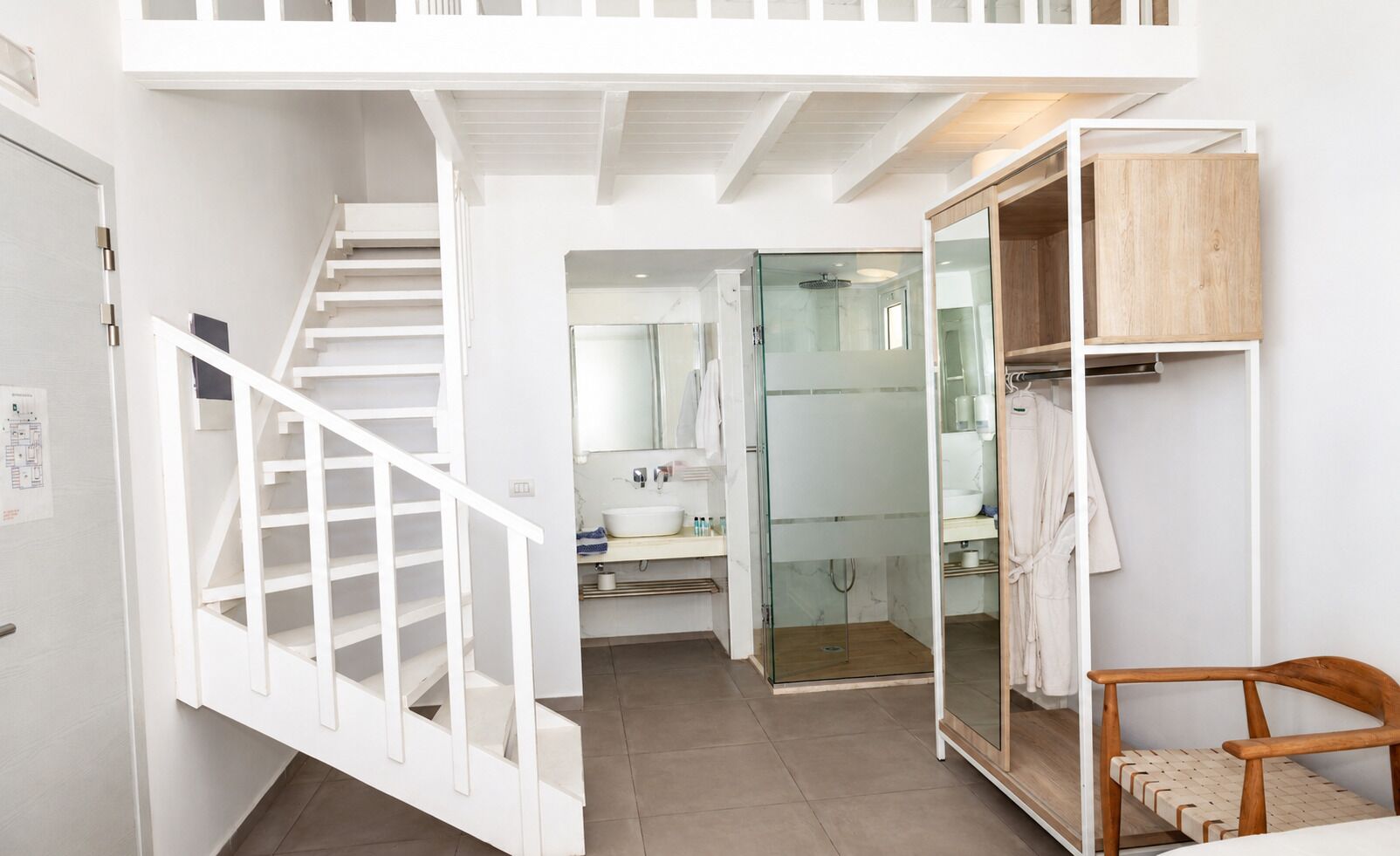 Un interior moderno con una escalera, una ducha con paredes de vidrio, una silla de madera y un armario blanco.
