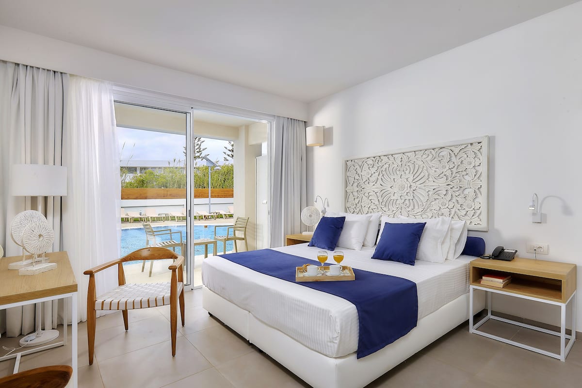 A hotel room with a large bed, a desk with a chair, a small table, and a view of a pool and outdoor seating.