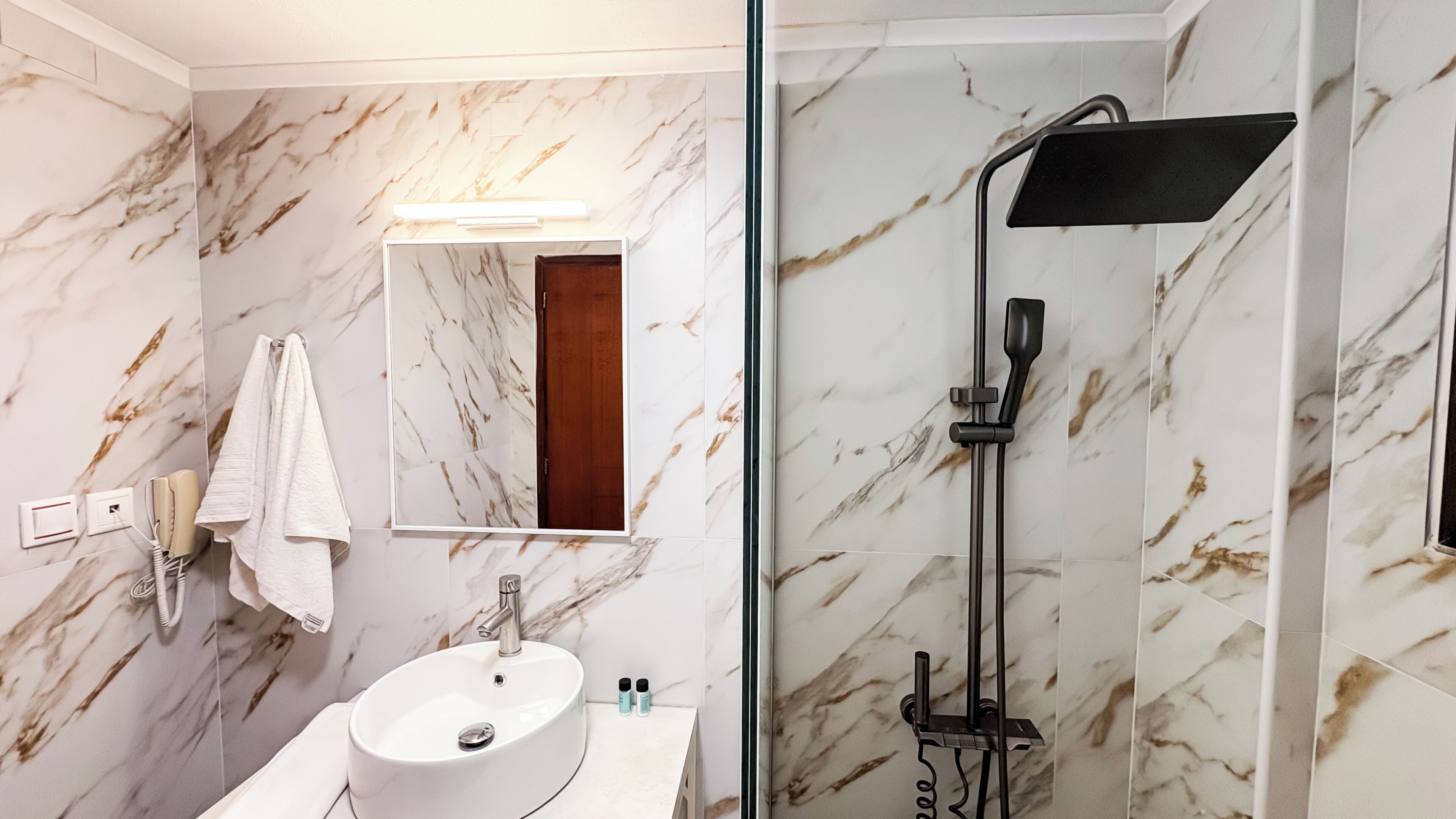 A bathroom with a marble wall, a sink with a mirror, a towel, and a shower with a handheld showerhead.