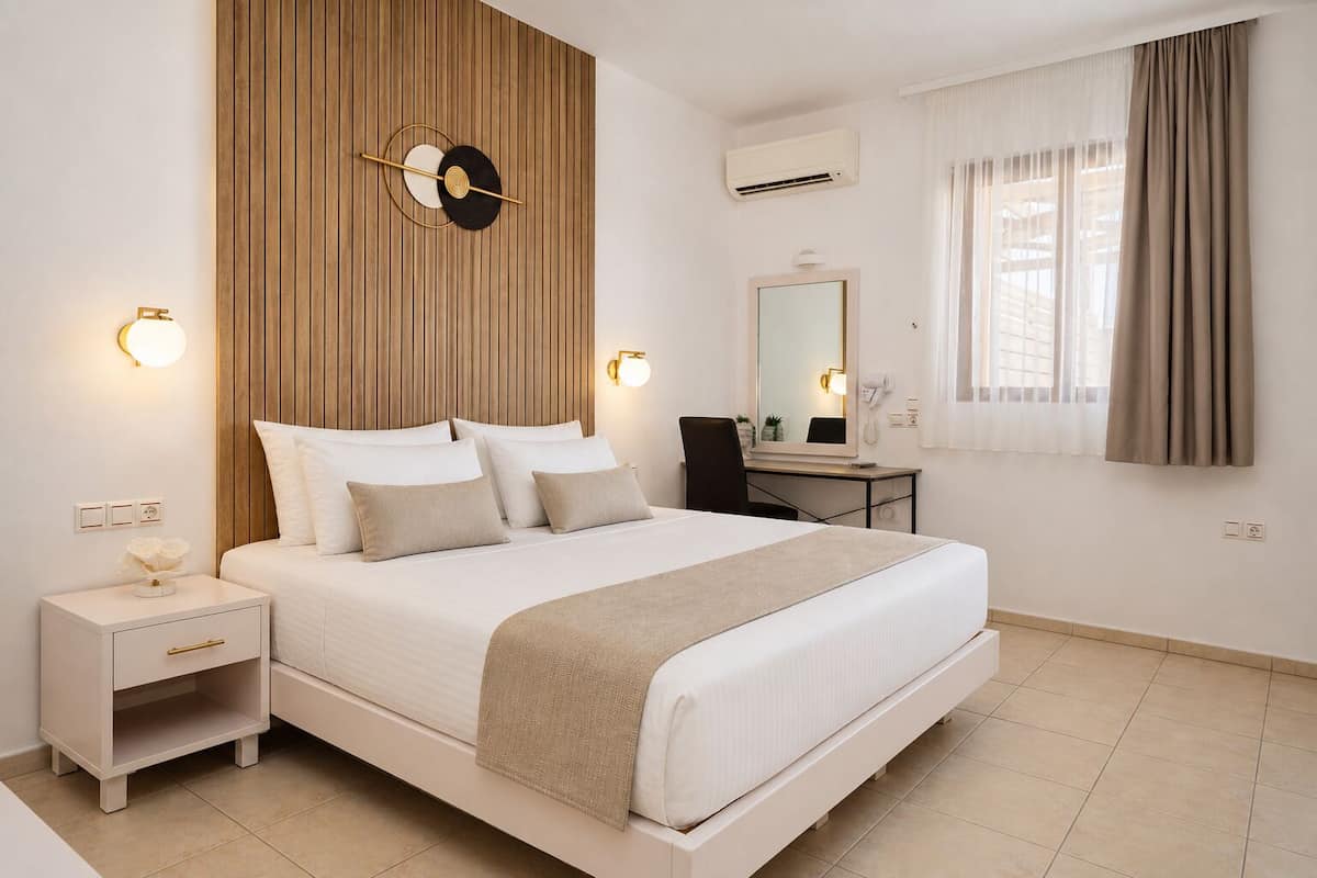 A hotel room with a bed, bedside table, lamp, mirror, and a view of the outside.